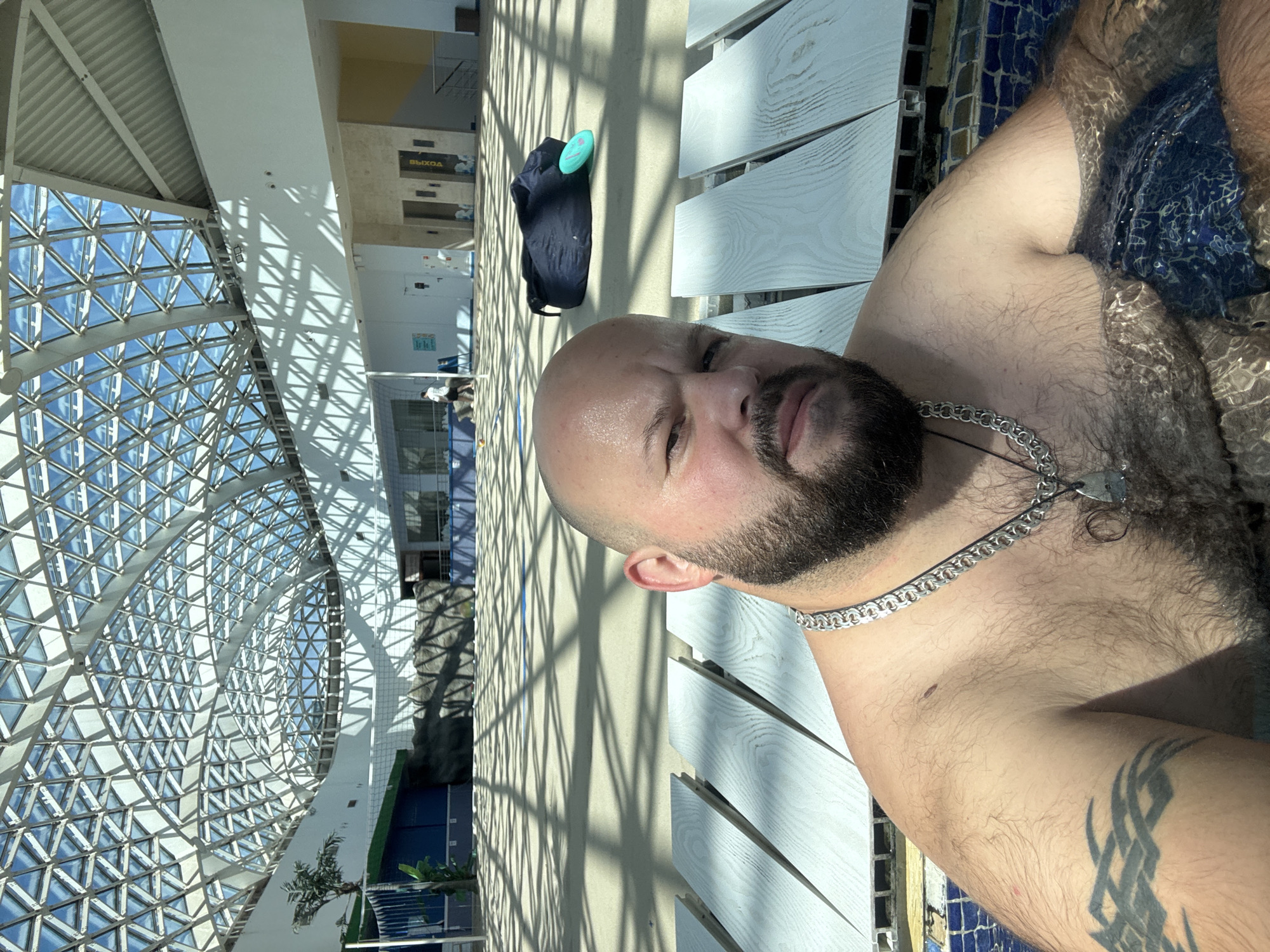 A man is in a hot tub at a water park in Estosadok, Russia. He is in the foreground, looking at the camera. He has a beard and a tattoo on his arm. The hot tub is surrounded by wooden benches. In the background, there is a large indoor space with a glass roof. There are lounge chairs and a beanbag chair on the floor. There are also elevators and stairs visible. The sun is shining through the glass roof, casting shadows on the floor.