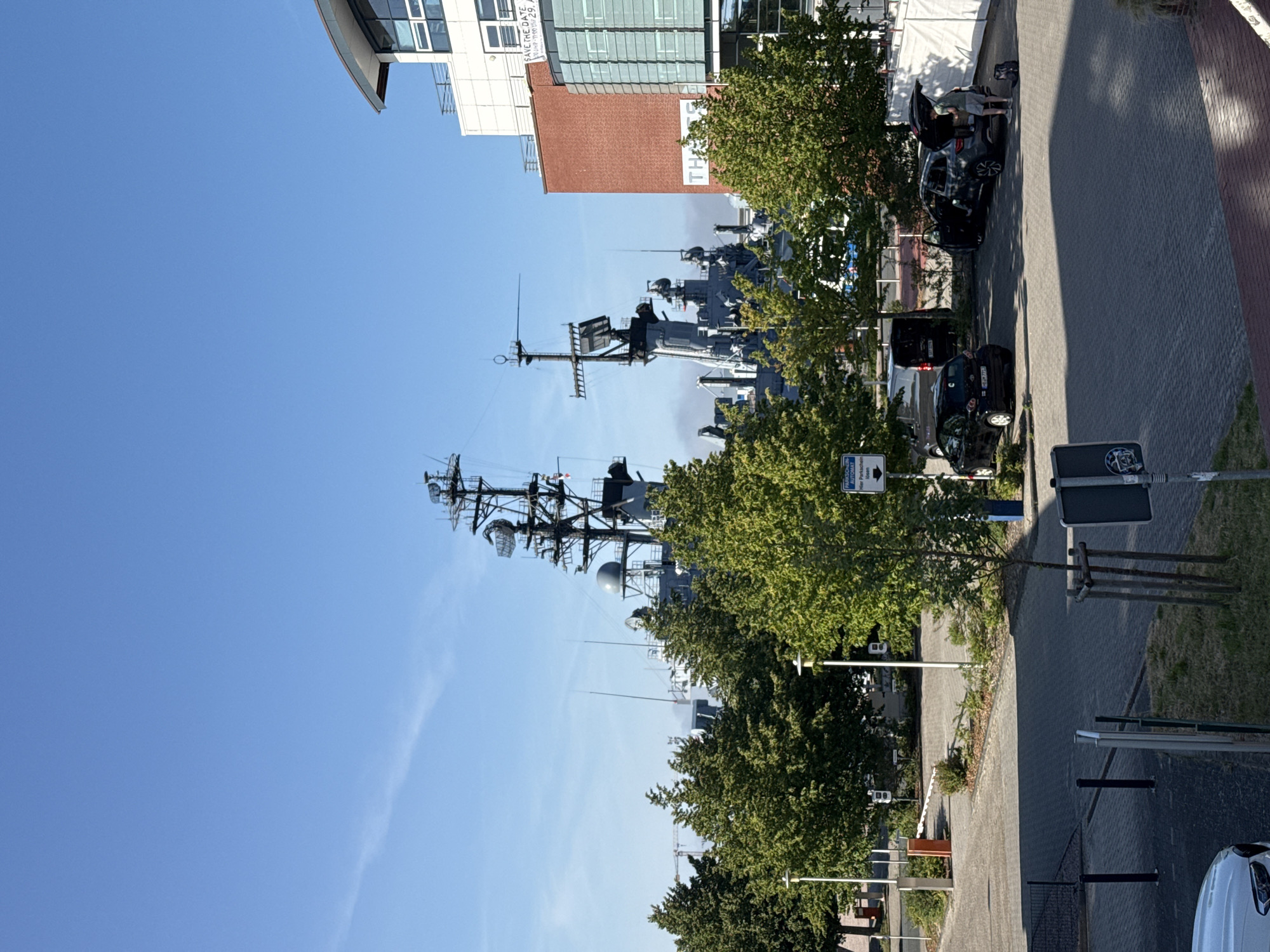 The image depicts a waterfront scene in Wilhelmshaven, Germany, with a large naval ship docked in the background. In the foreground, a paved parking lot is occupied by several vehicles, including a dark-colored hatchback and a grey SUV with its trunk open, where a person is seen interacting with the vehicle. Trees line the edge of the parking lot, separating it from a building with a distinctive red brick facade and large glass windows. The sky is clear and blue, suggesting fair weather. The overall scene captures a moment in a public area adjacent to a maritime facility.