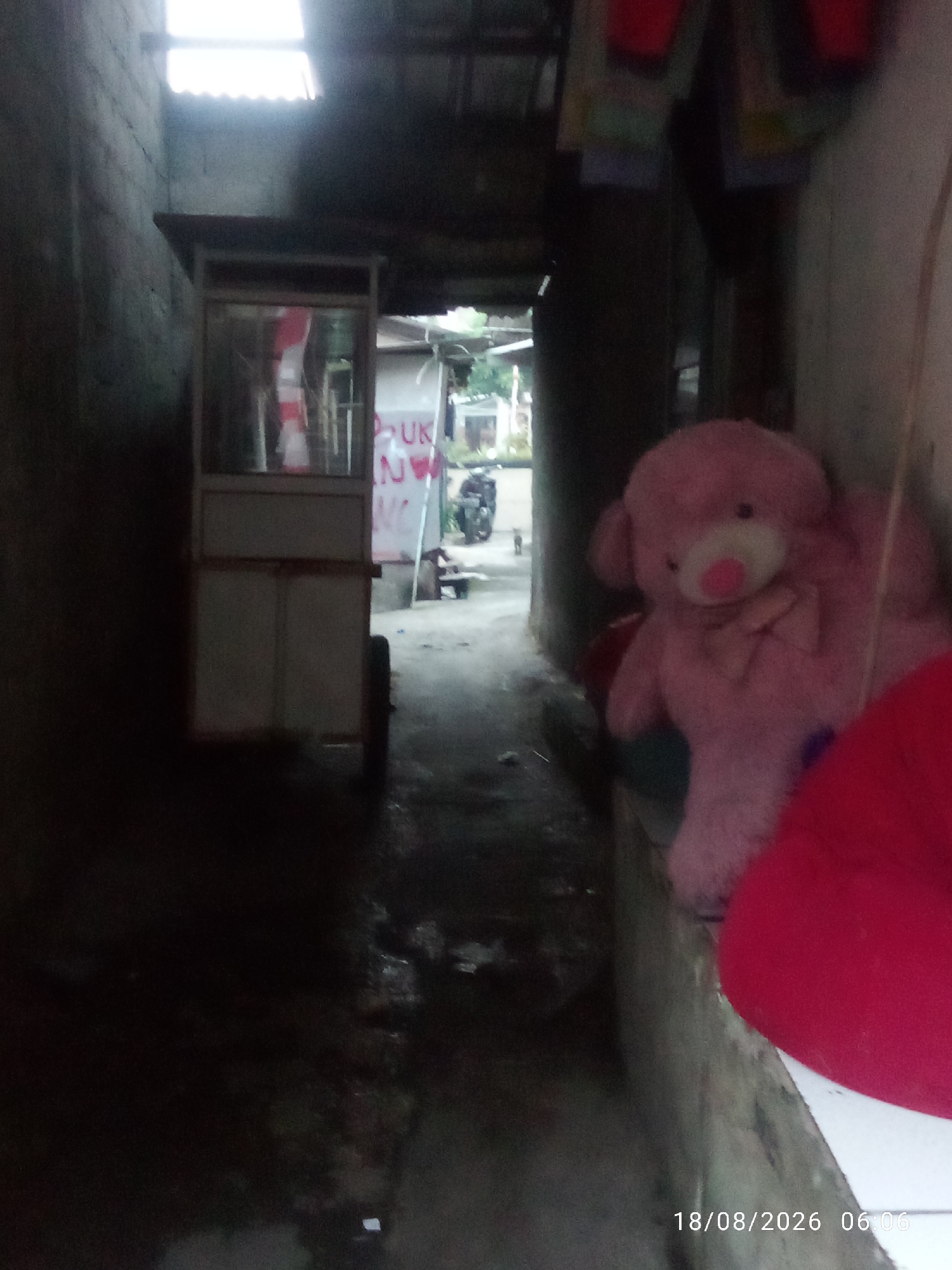 The image depicts a narrow alleyway, likely in East Jakarta, Indonesia, with a weathered, concrete wall on the left and a more textured, possibly plastered wall on the right. A small, mobile food cart with glass panels and white doors stands in the middle of the alley. Inside the cart, red and white striped objects are visible. Further down the alley, a large, soft, pink teddy bear is perched on a ledge or surface, with a bright red object, possibly a cushion, in the foreground and to the right. The alley floor appears wet and dirty. In the background, greenery and a motorcycle are faintly visible. The timestamp "18/08/2026 06:06" is present in the lower right corner.