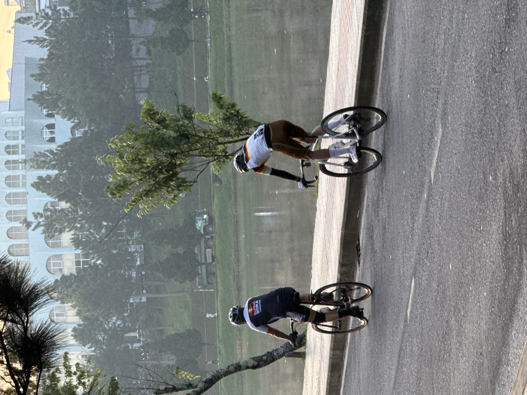 In Phường Xuân Hương - Đà Lạt, Vietnam, two individuals are cycling on a paved road adjacent to a body of water. The person in the foreground is wearing a dark cycling jersey with "BORA" in red lettering and a helmet. They are riding a bicycle with red rims. The second cyclist, further ahead, wears a white and yellow cycling jersey with German flag colors on the sleeve and brown cycling shorts. This cyclist is also wearing a helmet and white socks, and is on a bicycle with black rims. The background shows a misty landscape with trees, a light blue building with arched windows, and a green truck on a grassy area. The sun is rising, casting a warm glow on the scene.