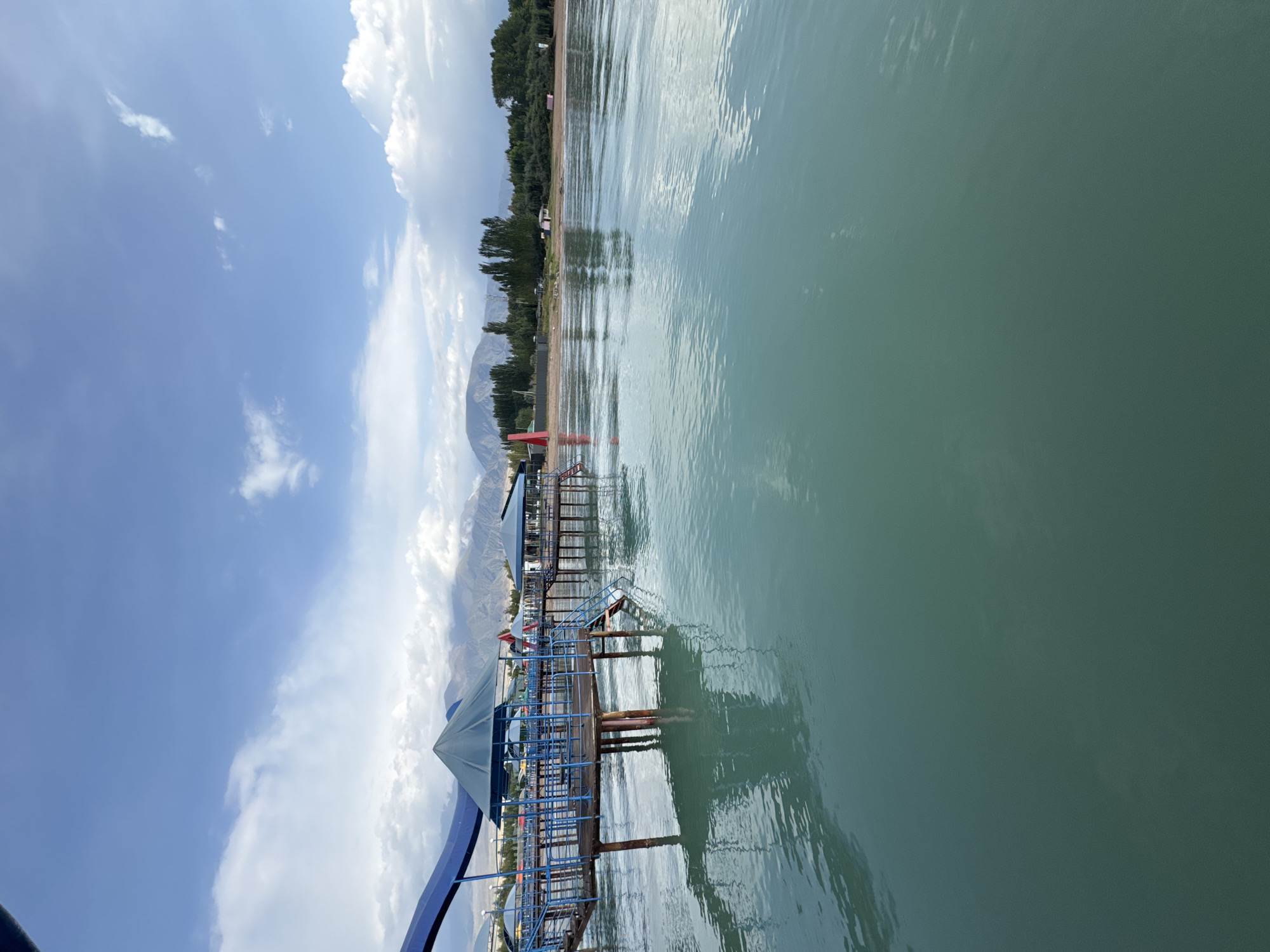 The image shows a waterfront scene in Sary-Oy, Kyrgyzstan. In the foreground, a body of calm, greenish-blue water fills the lower half of the frame, reflecting the sky and structures. A wooden pier with blue railings extends into the water from the left, supporting two covered gazebos. One gazebo is larger and closer, with a pyramidal roof, and a red slide is visible behind it. A second, smaller gazebo is visible further down the pier to the right. Steps lead down from the pier into the water. In the background, a sandy beach is lined with trees, and beyond that, a range of mountains with some snow-capped peaks is visible under a partly cloudy sky. The overall atmosphere is serene and suggests a recreational area.