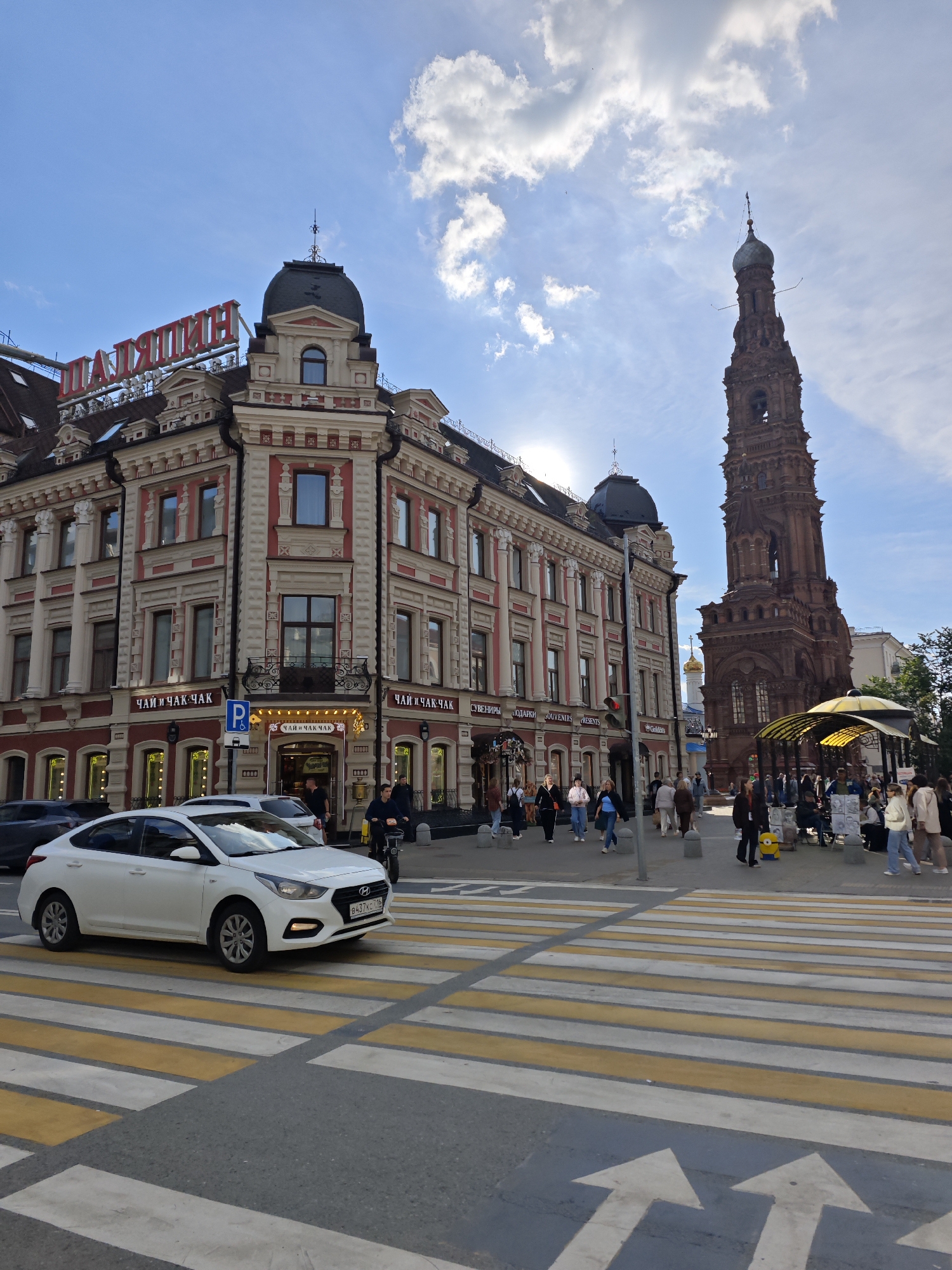 This is a photograph of a street scene in Kazan, Russia, likely taken on a sunny day with scattered clouds. In the foreground, a white Hyundai car is driving across a pedestrian crossing with yellow and white stripes. The street is paved, and directional arrows are visible on the road.
The background features a prominent, ornate building with red and beige accents, topped with a dark dome. A large red sign with Cyrillic lettering, reading "ШАЛЯПИН" (Shalyapin), is mounted on the building. Below this, the words "ЧАЙ И ЧАК ЧАК" (Tea and Chak-Chak) appear on several signs, indicating shops or cafes. One sign also includes the English text "SOUVENIRS PRESENTS".
To the right of the building, a tall, slender, reddish-brown tower, characteristic of a bell tower, rises against the sky. There are people walking on the sidewalk, some browsing at stalls, and a few vehicles, including a motorcycle. The presence of shops selling souvenirs and tea, along with the distinctive architecture, suggests a tourist area. The sun is bright, casting some shadows and illuminating the scene, giving the impression of it being midday or early afternoon.