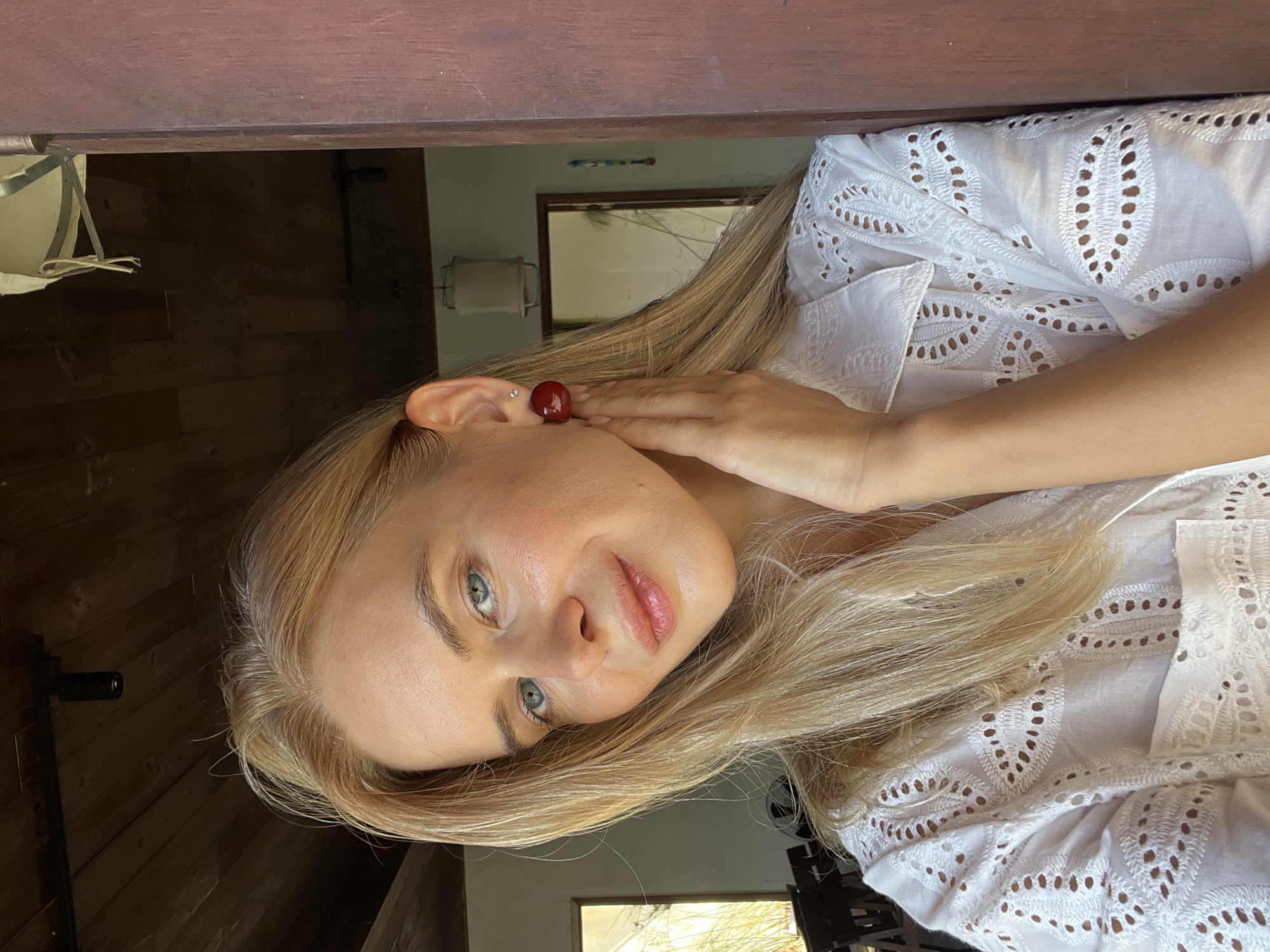The media file is a vertical, close-up, selfie-style portrait of a young Caucasian woman with blonde hair and blue-green eyes. She is wearing a white eyelet shirt. Her right ear is visible, adorned with a single cherry as an earring, and a small stud earring. Her hand is gently touching her face near her ear. The woman is looking directly at the camera with a soft, relaxed expression. The lighting suggests it is daytime, likely indoors or in a shaded outdoor area, given the soft light on her face and the reflections in the background.
The background is dimly lit and appears to be an interior space with wooden walls. In the background, a mirror reflects a window or doorway with a view of greenery, possibly palm trees, which, combined with the known location context of Nam Nha Trang Ward, Vietnam, suggests a tropical or sub-tropical setting. The overall impression is one of a casual, intimate moment captured by the subject herself. There is no visible text in the image.