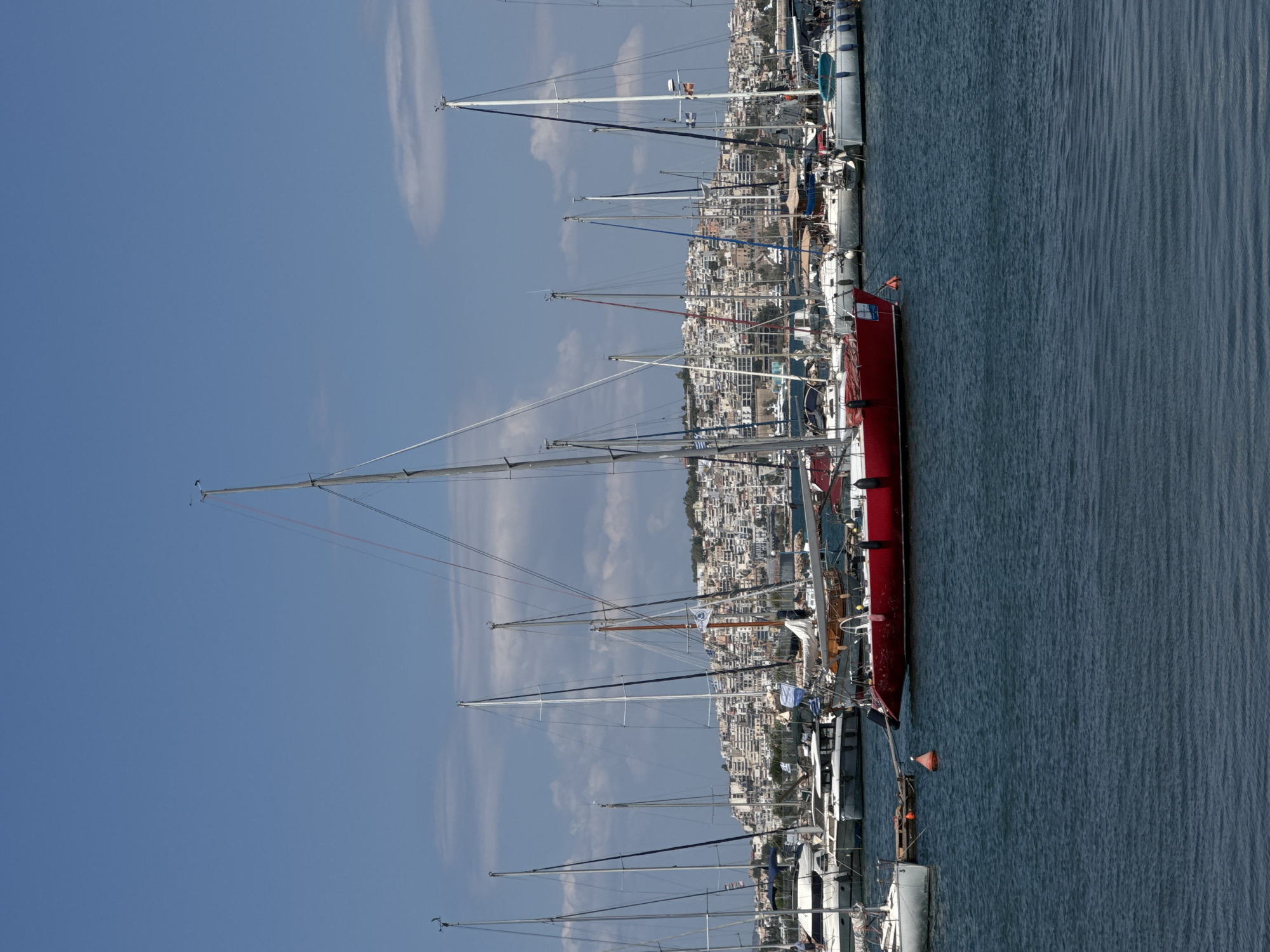 This is a photograph of a harbor in Athens, Greece, on a clear day with a blue sky and scattered clouds. In the foreground, several sailboats are docked on dark blue water. The most prominent vessel is a bright red sailboat in the center of the frame, positioned slightly to the left. It has a white cabin and mast. To the left of the red sailboat is a white sailboat tied to a wooden dock. In the background, a dense cityscape of white buildings with red-tiled roofs climbs a hill, characteristic of Athens. The harbor is filled with numerous other sailboats of various sizes and colors, their masts creating a dense forest of rigging against the sky. Some boats are white, others are darker, and some have visible sails. A few small flags are visible on some of the boat masts. The overall impression is a busy, sunny day at a Greek marina with the urban sprawl of Athens visible behind it.