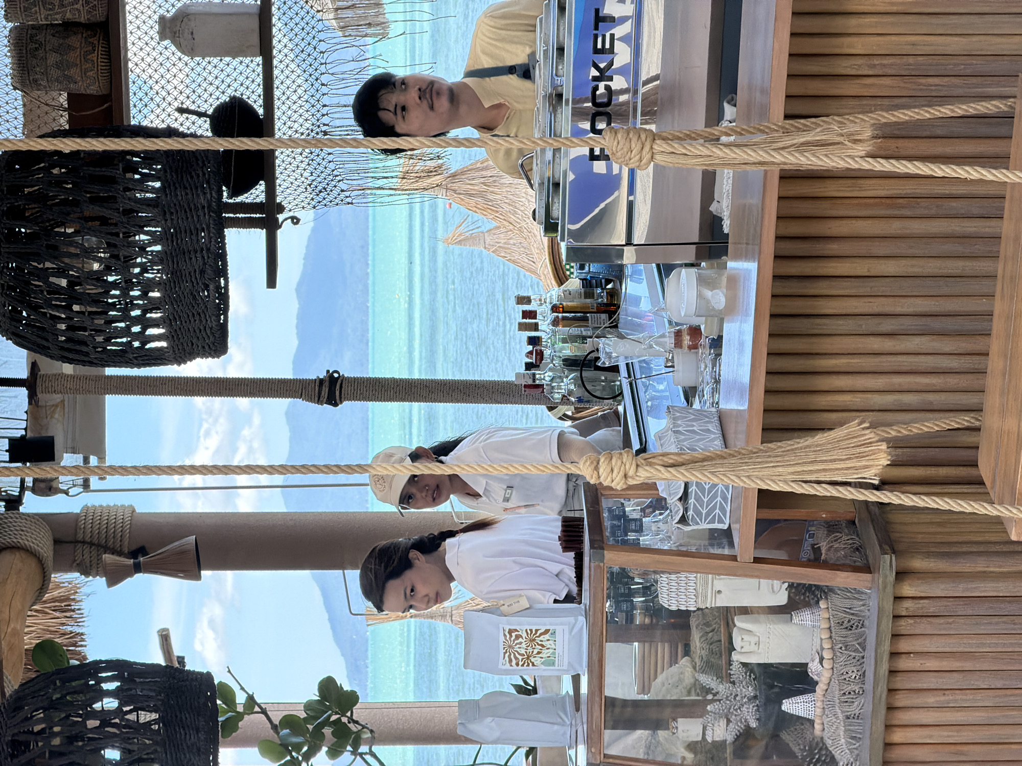 At a beachside cafe in Baan Bo Phut, Thailand, three individuals are present behind a wooden counter. A young woman with her hair in a braid stands to the left, looking away from the camera. Next to her, a woman wearing a cap and a white polo shirt is looking forward. To the right, a man with a mustache and dark hair, wearing a light yellow apron over a beige shirt, operates a chrome espresso machine with "ROCKET" visible on its side. Bottles of alcohol are lined up behind the bar, and various decorative items, including woven baskets and ceramic figures, are displayed on shelves and within glass cases. The cafe overlooks a bright blue sea and distant, hazy mountains under a clear sky. Ropes hang from the ceiling, one of which is knotted and forms part of a swing structure. The overall ambiance is relaxed and tropical.