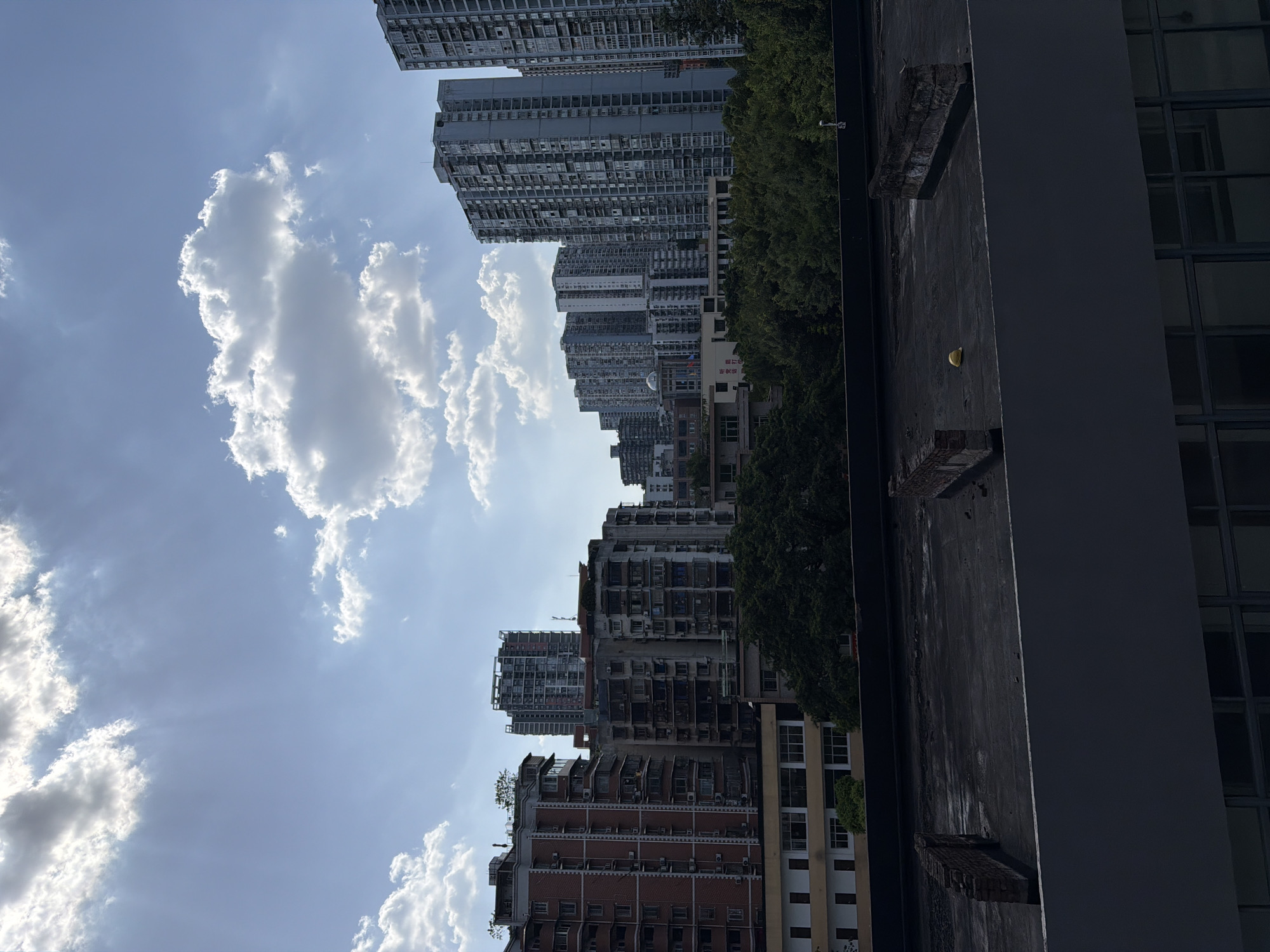 The image depicts a cityscape, likely in Jiulongpo District, China, under a partly cloudy sky with visible sunbeams. In the foreground, a dark, flat rooftop is visible, with some weathered brick structures and debris. Beyond the rooftop, a cluster of multi-story apartment buildings of varying architectural styles and ages are densely packed together. Some buildings are constructed with red brick, while others are taller, modern concrete structures with numerous windows. Lush green trees are interspersed between and behind some of the buildings. A yellow hard hat is present on the rooftop in the middle ground. The overall scene conveys an urban environment with a mix of residential and possibly commercial structures, set against a backdrop of a bright, dynamic sky.