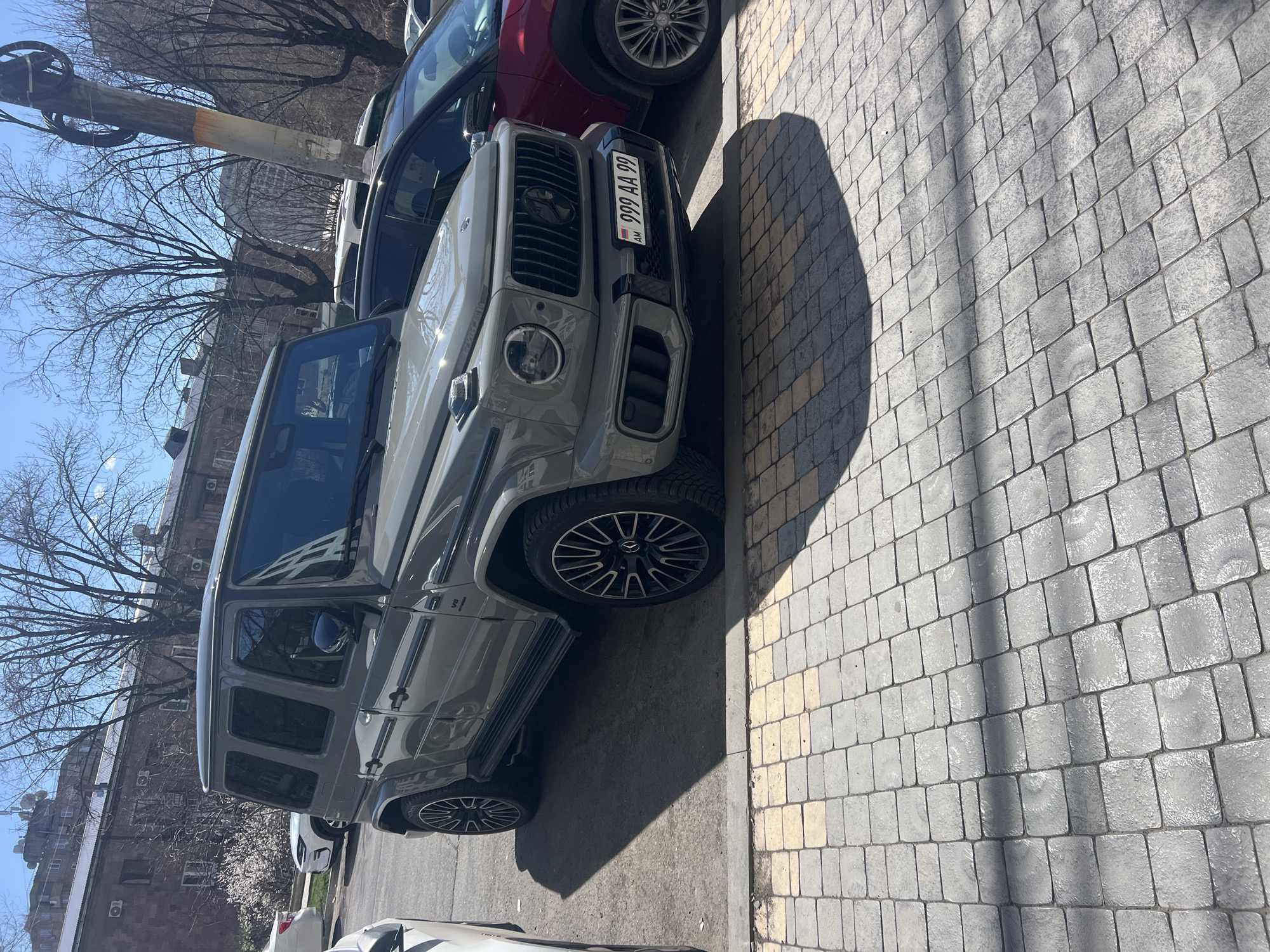 The image shows a gray Mercedes-Benz G-Class parked on a street in Yerevan, Armenia. The car's license plate reads "999 AA 99". The street is paved with gray stone blocks. Other vehicles are visible in the background, including a red car. Bare trees line the street, and buildings are visible in the background. The sky is blue.
