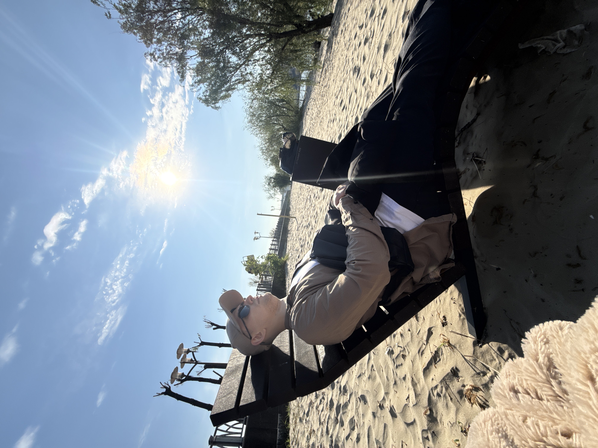 A man lies on a park bench on a sandy area in Rostov-on-Don, Russia. The man is wearing a beige cap, sunglasses, a beige shirt, a black backpack, and black pants. He is lying on his back, looking up at the sky. The park bench is made of dark wood. The sandy area is covered with a light-colored sand. In the background, there are trees and a blue sky with clouds and the sun. The lamp posts with pruned trees are also visible.