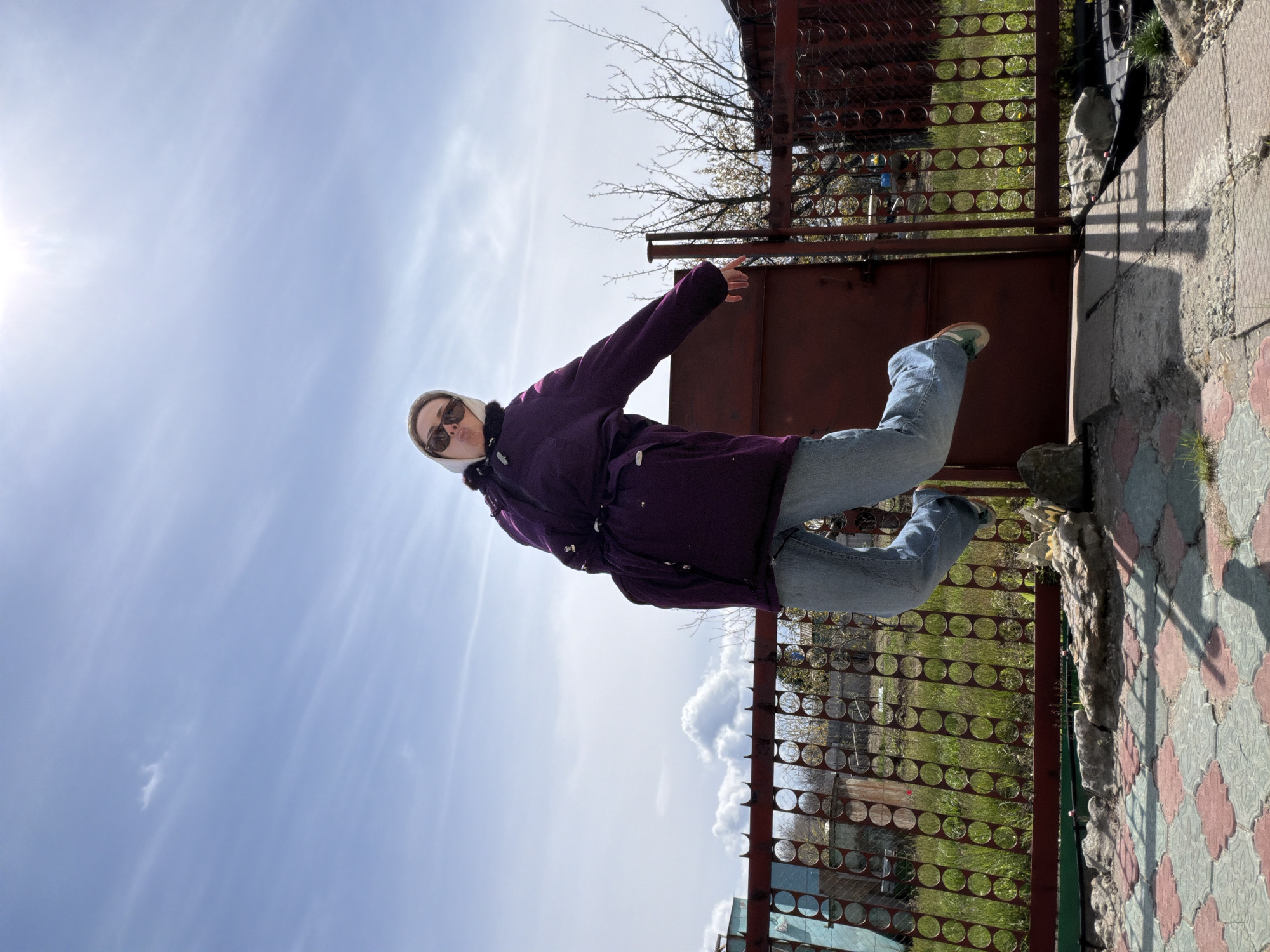 A woman wearing a purple winter coat, blue jeans, a white hat, and sunglasses is standing near a gate in Kamyshin, Russia. The gate is made of brown metal with a circular pattern. A stone path is visible on the ground, and beyond the gate, there is green grass and other vegetation. The sky is blue with some clouds, and the sun is shining.