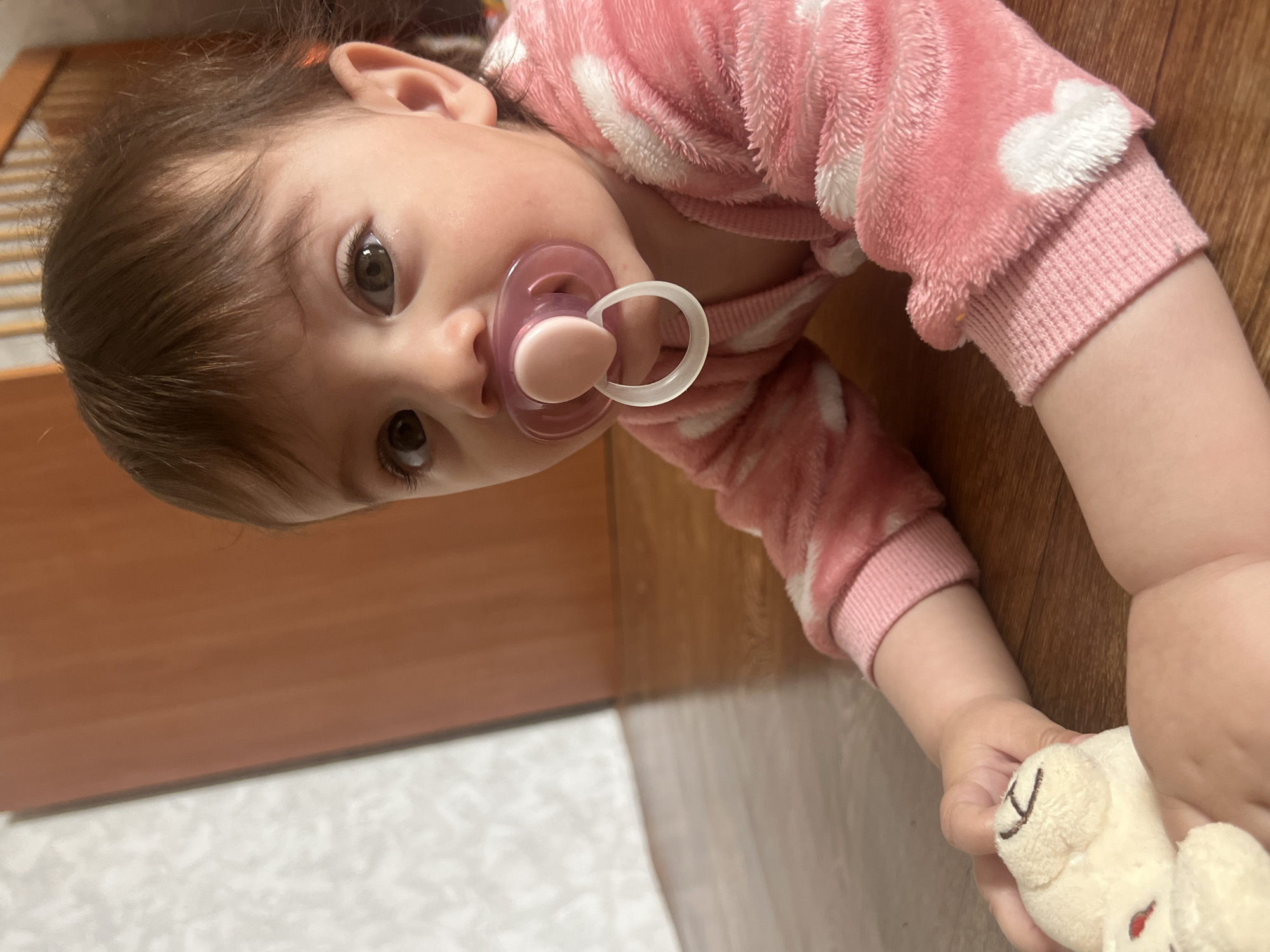 A baby with brown hair and eyes lies on a wooden floor. She is wearing a pink and white polka-dotted onesie and has a pink pacifier in her mouth. She holds a small, white stuffed animal in her right hand. The image does not contain sufficient information to determine the location in the city of Elena, Bulgaria.