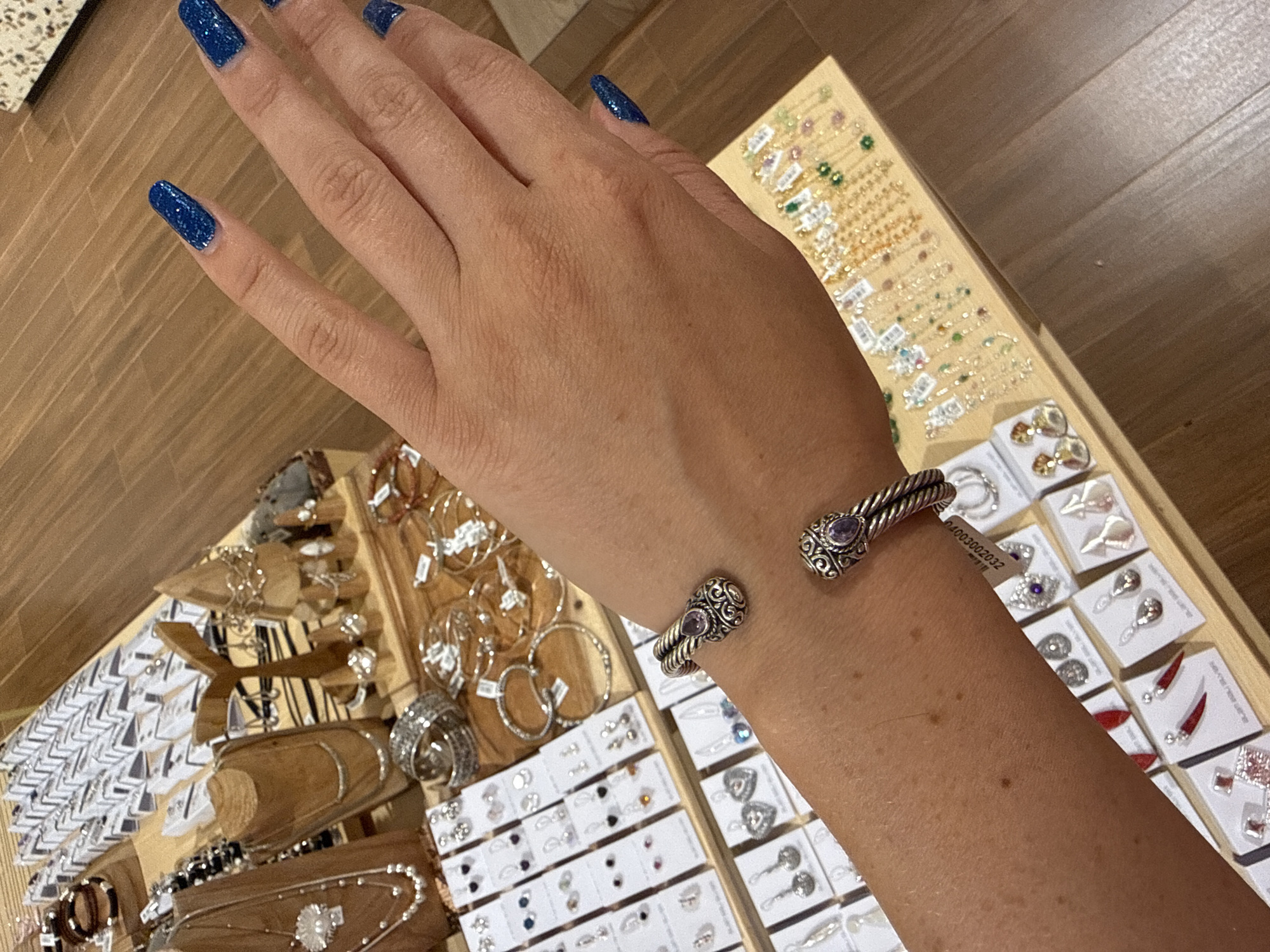 A person's right forearm and hand are visible in the foreground, adorned with a silver cuff bracelet featuring a purple stone. The bracelet is positioned on the forearm. The hand is partially visible, with fingers extended. The nails are painted a vibrant blue glitter.
In the background, a display of jewelry is organized on wooden tables. Various types of jewelry, including necklaces, bracelets, and earrings, are presented in rows on small white cards. Some necklaces appear to be delicate chains with colorful gemstones. Other displays showcase silver-toned jewelry, including chunky bracelets and intricate earrings. The overall environment suggests a retail setting, possibly a jewelry store or market stall. The flooring is made of wood planks.
The location is identified as Unknown, Indonesia.