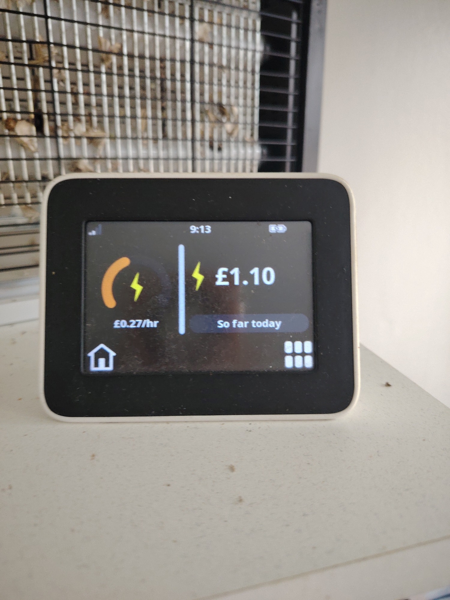 An upward-angled, eye-level indoor shot depicts a white smart energy meter with a black screen positioned on a light-colored, soiled surface in Huntingdonshire, United Kingdom. The meter's screen displays "9:13" at the top center. On the left side, an orange curved bar with a yellow lightning bolt indicates a current cost of "£0.27/hr," alongside a home icon at the bottom left. On the right, a yellow lightning bolt preceding "£1.10" shows the cost "So far today," with a grid icon at the bottom right. A signal strength icon is visible top left, and a battery icon top right. The screen itself is visibly dusty and smudged. The light-colored surface beneath the meter is also covered in small specks of dirt and dust. In the blurred background to the left, a metallic grid structure with scattered brownish debris is partially visible. To the right background, a dark, possibly louvered or vented structure is seen next to a plain, light-colored wall.