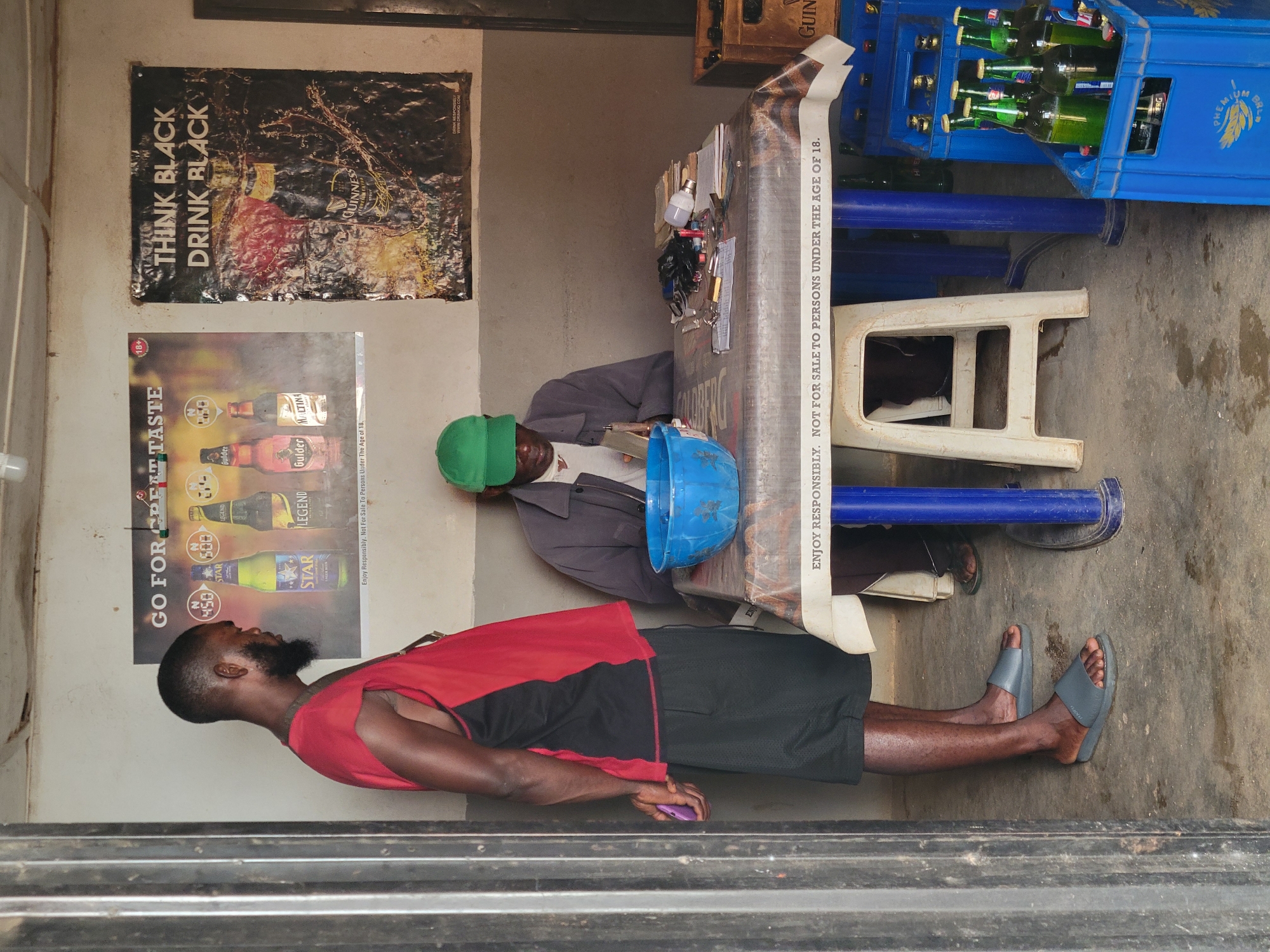 In Barakin Lerawi, Nigeria, two men are present within an indoor establishment. One man, standing on the left, faces away from the camera. He has dark skin, a beard, and is wearing a red and black sleeveless top and black shorts. He holds a purple object in his left hand. The other man sits at a table on the right, facing the camera, wearing a gray jacket and a green cap. He appears to be interacting with items on the table, which is covered with a patterned tablecloth. Various objects are on the table, including a blue bowl, what appears to be a stack of papers, writing utensils, a lightbulb, and other small items. Behind the man at the table, a wooden crate is visible. In front of the table, to the right, a blue plastic crate filled with green glass bottles is present, and another similar crate is visible to the left, partially obscured by the table. Posters are displayed on the wall behind the men. The floor is concrete, with some dampness.