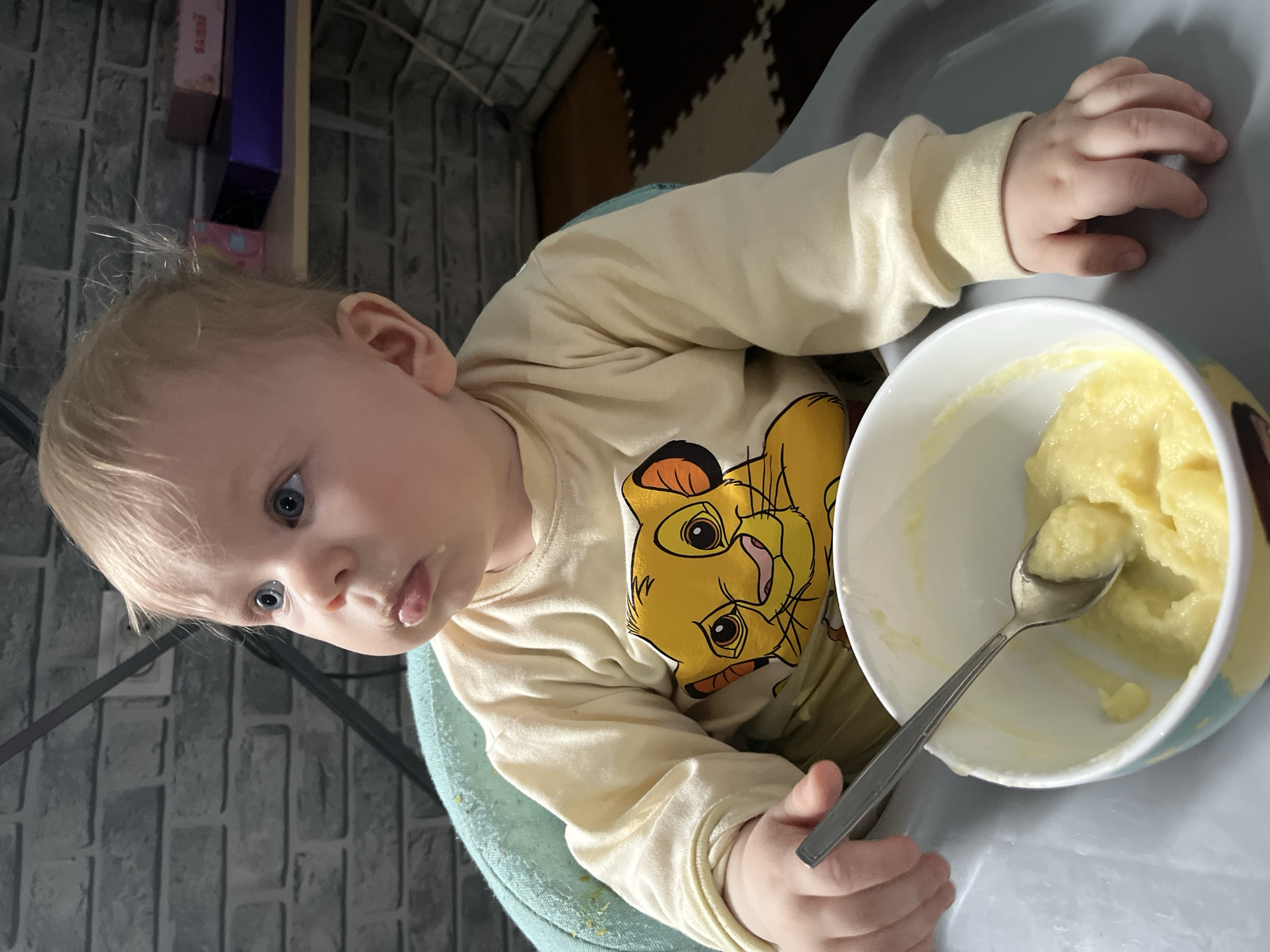 A baby with blond hair and blue eyes, wearing a yellow shirt with a Simba cartoon character on it, sits in a high chair. A white bowl containing mashed food sits in front of the baby, with a spoon inside the bowl. Some of the food is on the baby's face. The background includes a grey brick wall and a brown and white patterned rug. There is no evidence of the image being taken in Vesyoloye, Russia.