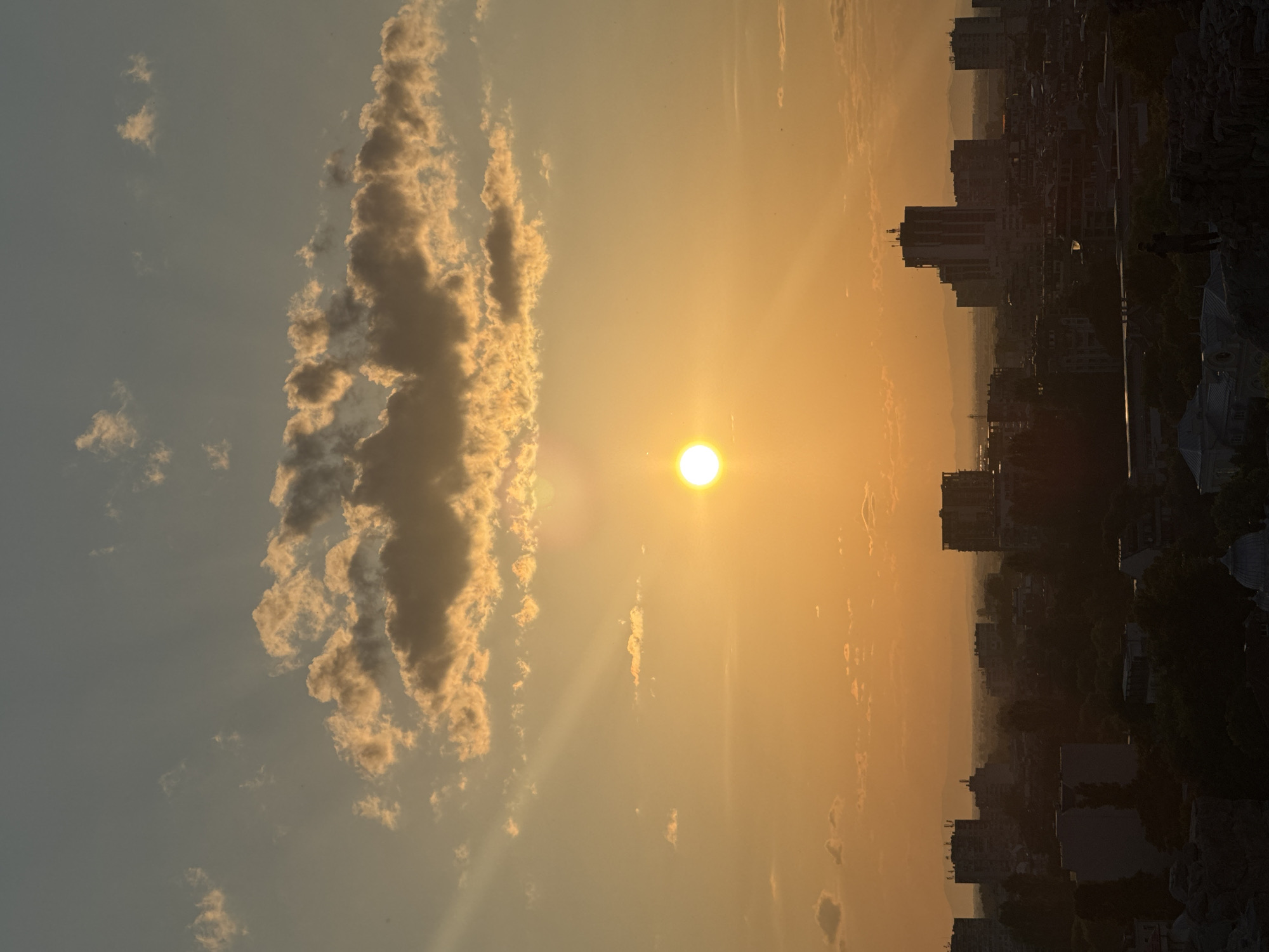 The image depicts a sunset over the city of Plovdiv, Bulgaria, viewed from an elevated position, likely one of its historic hills (e.g., Nebet Tepe). The sky is dominated by warm orange and yellow hues emanating from the bright, circular sun, positioned slightly right of center. Visible crepuscular rays extend from the sun. Scattered clouds are present, with those closer to the horizon illuminated in gold and those higher up appearing in darker silhouette against a muted blue-grey upper sky.
The mid-ground reveals a sprawling urban landscape of Plovdiv, with numerous multi-story buildings and structures rendered in dark silhouette against the vibrant sky. The details of individual buildings are obscured by the low light and atmospheric conditions, but varying heights and densities of construction are apparent. Distant mountains or hills are faintly visible on the horizon, softened by atmospheric haze.
In the foreground, dark, rugged rock formations or remnants of ancient stone structures are visible, consistent with a historical viewpoint. A single human figure stands in silhouette on a rocky outcrop or wall in the lower right quadrant, facing the sunset and the city below. Partial views of architectural features, including a light-colored domed structure, are discernible at the very bottom of the frame. The overall scene captures the dramatic light of a late afternoon or early evening in an urban setting.