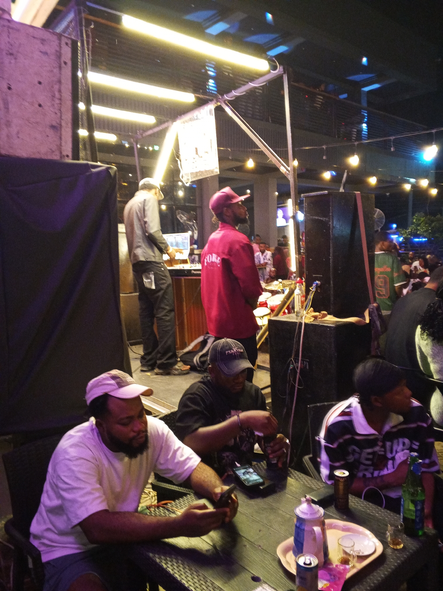 The image depicts a scene in what appears to be an outdoor bar or restaurant setting, likely in Lokoja, Nigeria. Four men are seated at a dark-colored table. One man wears a white t-shirt and cap, looking at a phone. Another man is in a dark t-shirt with a cap, seemingly preparing to smoke or use a pipe. A third man wears a striped shirt and cap, looking away from the table. On the table are cans of soda, a bottle of what appears to be beer, a tray with cups, a tea kettle, and a cell phone. Behind them, a man in a red jacket is standing, and another man in a gray shirt is operating a laptop on a DJ console. Audio speakers and lighting equipment suggest a performance or DJ setup. Additional people are visible in the background seated at other tables. The area is lit by overhead string lights and fluorescent tubes.