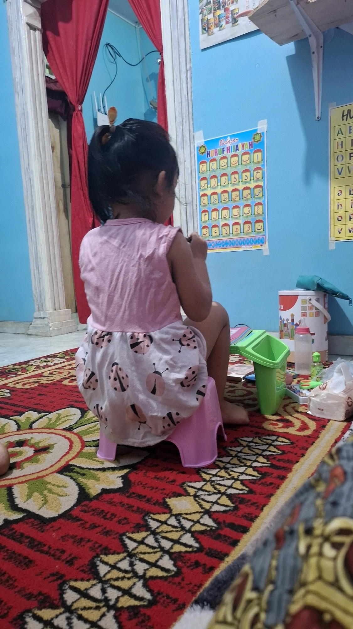 A young child, viewed from behind, sits on a pink potty chair in what appears to be a child's bedroom or playroom. The child is wearing a pink sleeveless shirt and a light-colored skirt adorned with ladybug prints. The room has bright blue walls. A poster with colorful letters is affixed to the wall to the right of the child. Various toys and supplies are scattered on a red rug with a patterned design. Red curtains hang in the background, framing a doorway. The overall impression is of a domestic setting where a child is engaged in potty training or playing.