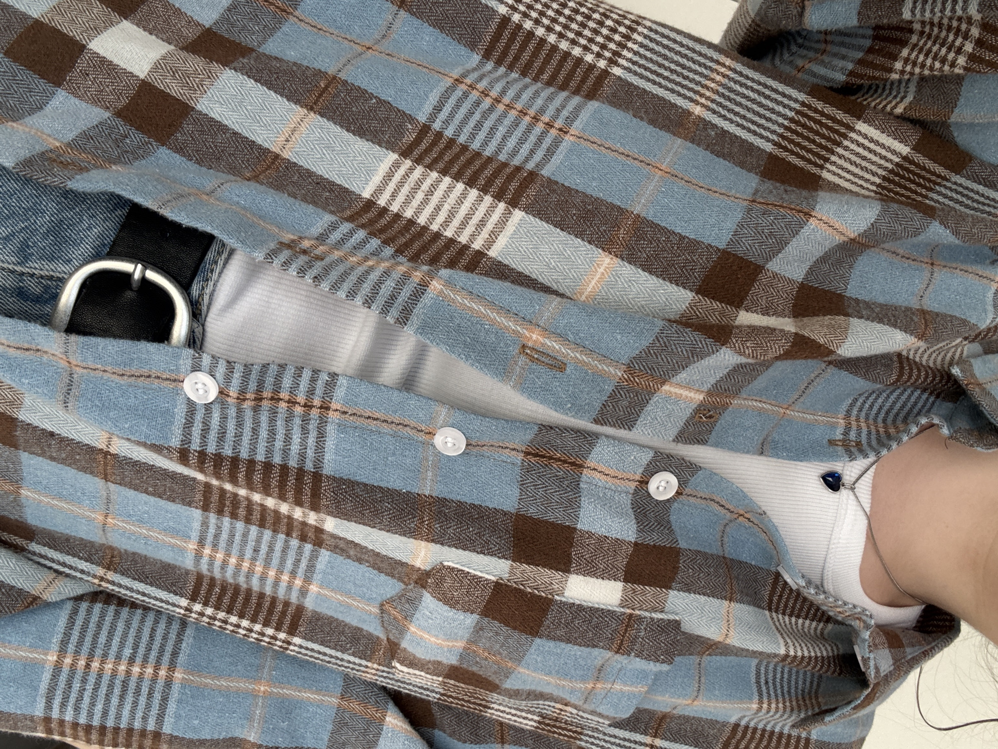This is a close-up, chest-up shot of a person wearing a light blue, brown, and white plaid flannel shirt over a white ribbed tank top. A silver chain with a blue heart-shaped pendant is visible around the person's neck. The shirt has white buttons down the front. The lower part of the image shows a black belt with a silver buckle over blue denim jeans. The background is a plain, light-colored surface. There is no information available about the location of this image in Lyubertsy, Russia.