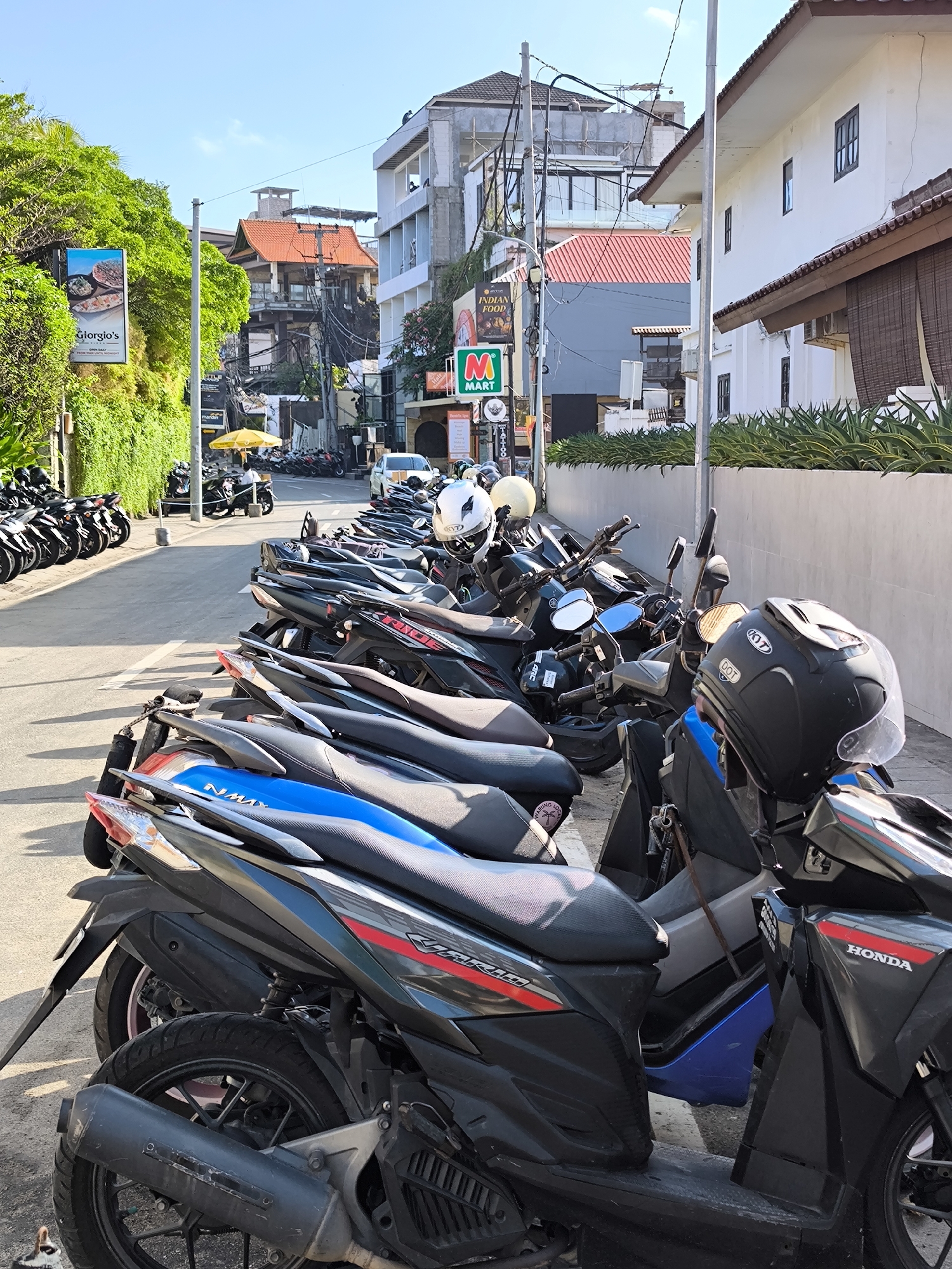 The image captures a sunny day in Tibubeneng, Indonesia, showcasing a street densely packed with parked scooters. The foreground is dominated by rows of scooters, predominantly black and blue, with visible Honda branding on some. Many have helmets resting on their seats.
In the background, various buildings line the street, including a white, two-story structure with a brown-tiled roof and a gray building with a red-tiled roof. Signage indicates the presence of restaurants and businesses like "M Mart" and "Giorgio's." The street scene suggests a bustling local environment common in Indonesian urban areas. A few people are visible in the distance near a yellow umbrella. A white car is parked further down the street.