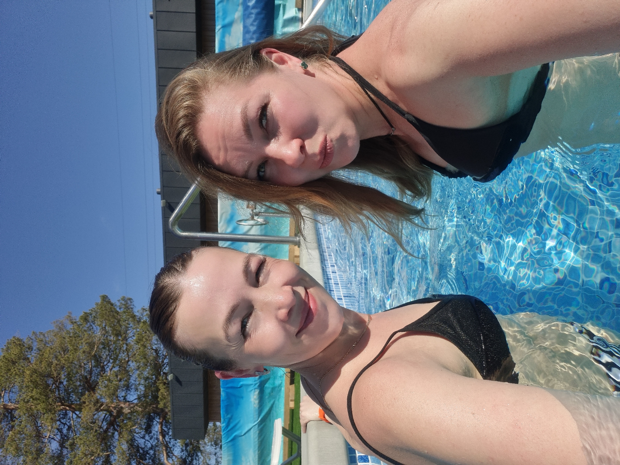 Two women are in a swimming pool with blue tile. Both are wearing black bathing suits and necklaces. The woman on the right is puckering her lips and looking at the camera. The woman on the left is winking and smiling. The sky is blue, and there is a building in the background. The location is Votkinsk, Russia.