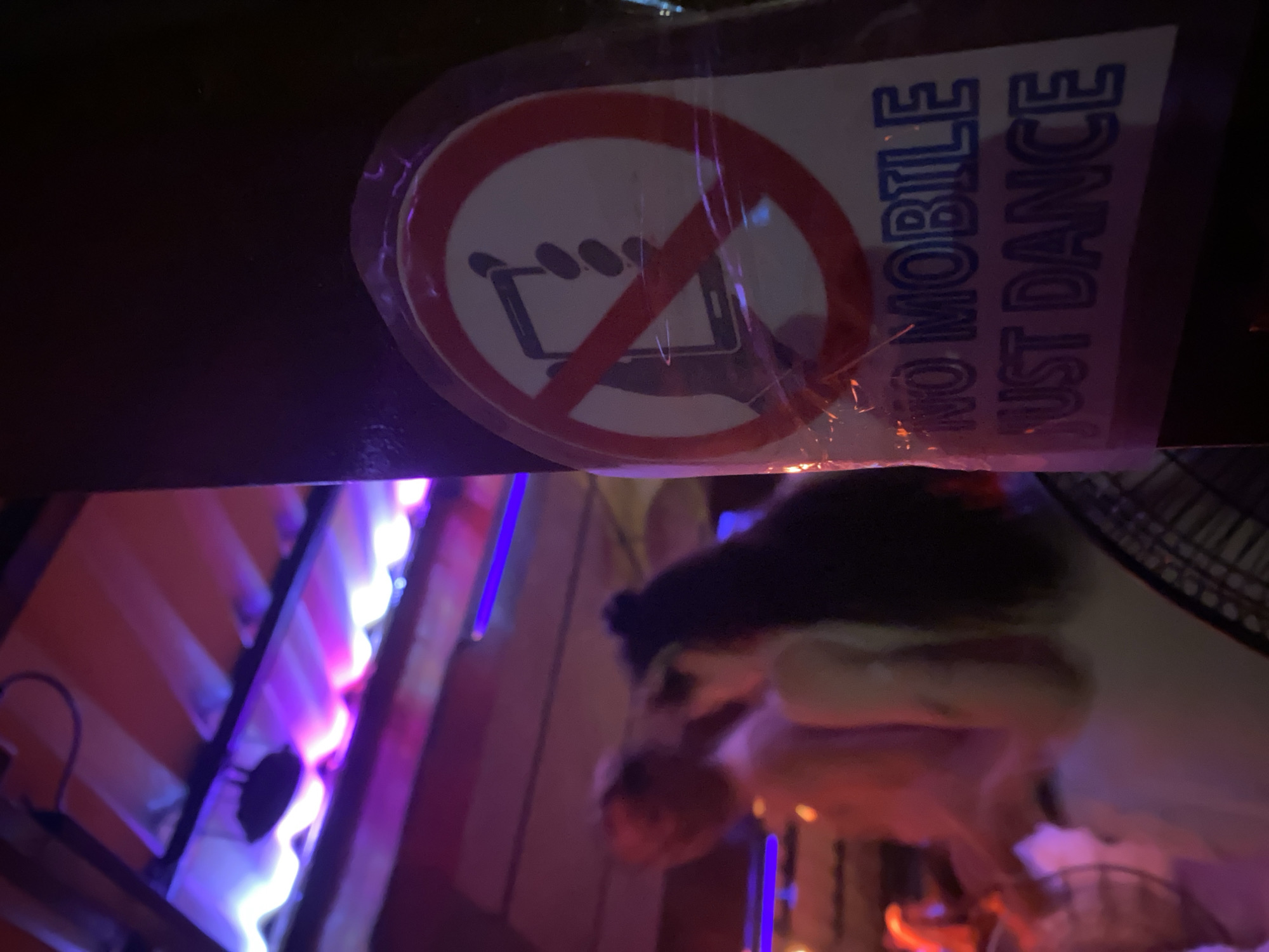 The image, taken indoors, possibly in a bar or club, features a sign that prohibits cell phone use. The sign shows a mobile phone inside a red circle with a line through it, and the text "NO MOBILE JUST DANCE". The location may be in ตำบลเกาะพะงัน, Thailand. The background is blurry, showing neon lights and possibly people. The composition suggests the image was taken from below, looking up at the sign.