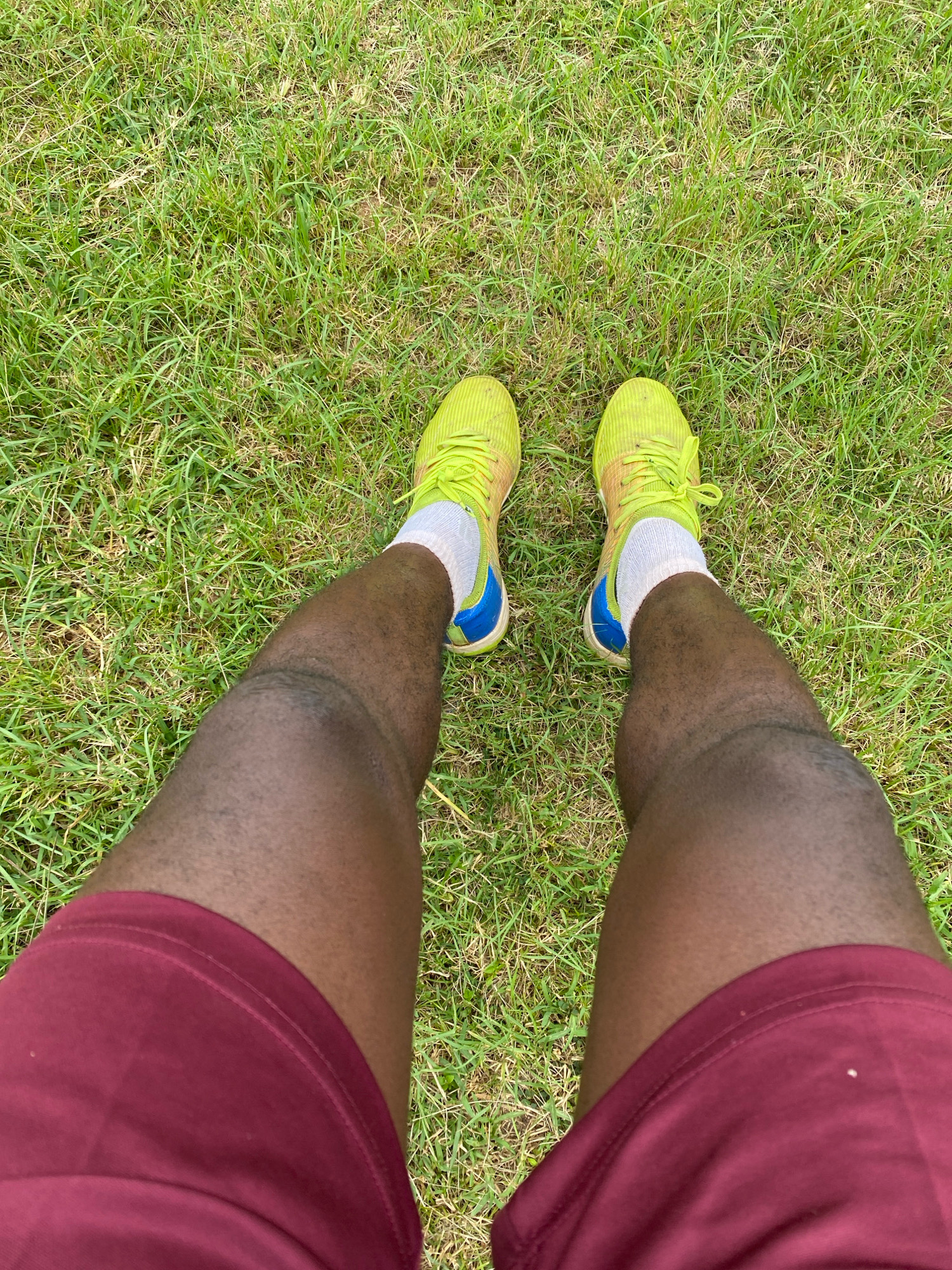 A person's legs are shown from the knees down, standing on a grassy field. The person is wearing maroon shorts and bright yellow athletic shoes with white socks. The shoes have blue accents. The grass is green and appears to be well-maintained. The image is shot from a high angle, looking down at the person's legs. No specific location in Jos, Nigeria can be identified from the image.