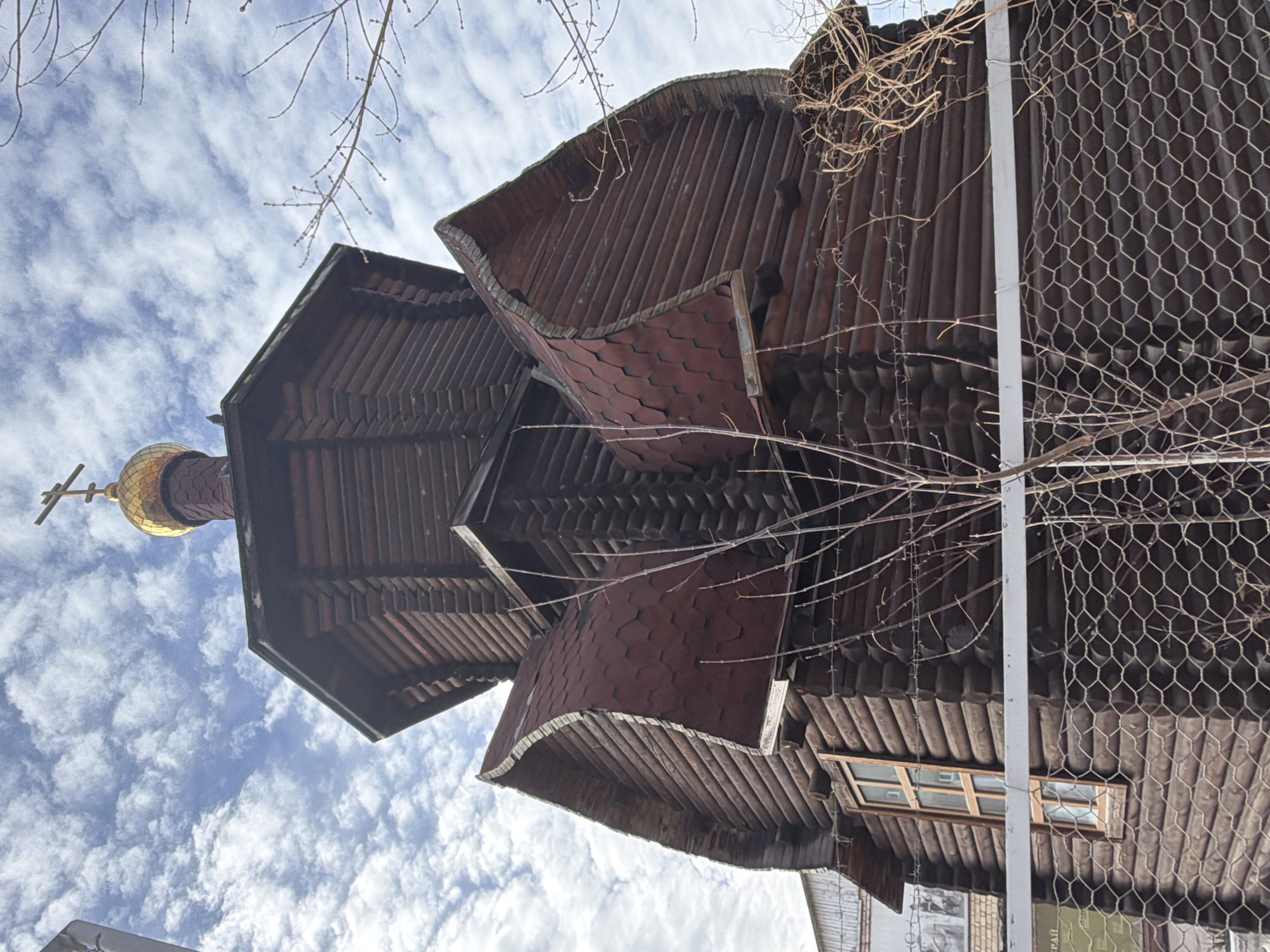Low-angle view of the St. Paraskeva Pyatnitsa Chapel in Krasnoyarsk, Russia, showing its tiered wooden construction and golden dome topped with a cross. The structure features distinct levels with overlapping roofs, some covered with dark red shingles. A window is visible on the side. A metal fence and bare tree branches are in the foreground. The sky is visible in the background.
