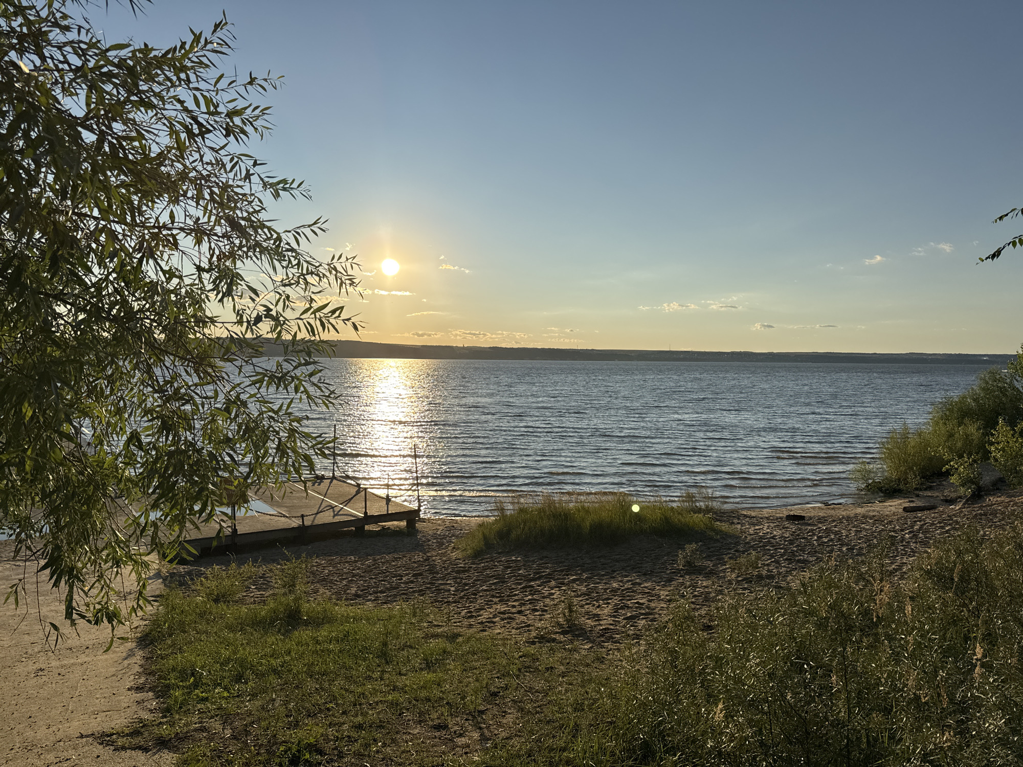 A serene sunset over a wide body of water, possibly a river or lake, is depicted. The sun, a bright orb, casts a shimmering reflection across the water's surface, creating a path of light. The sky transitions from a clear blue at the top to a warm, golden hue near the horizon, with a few wispy clouds. The foreground features a sandy beach dotted with patches of green grass and foliage. On the left, the branches of a leafy tree hang over the scene, framing the view. A small wooden pier juts out into the water from the beach. The environment appears natural and undisturbed, suggesting a peaceful, outdoor setting. The location is identified as Matyushino, Russia.