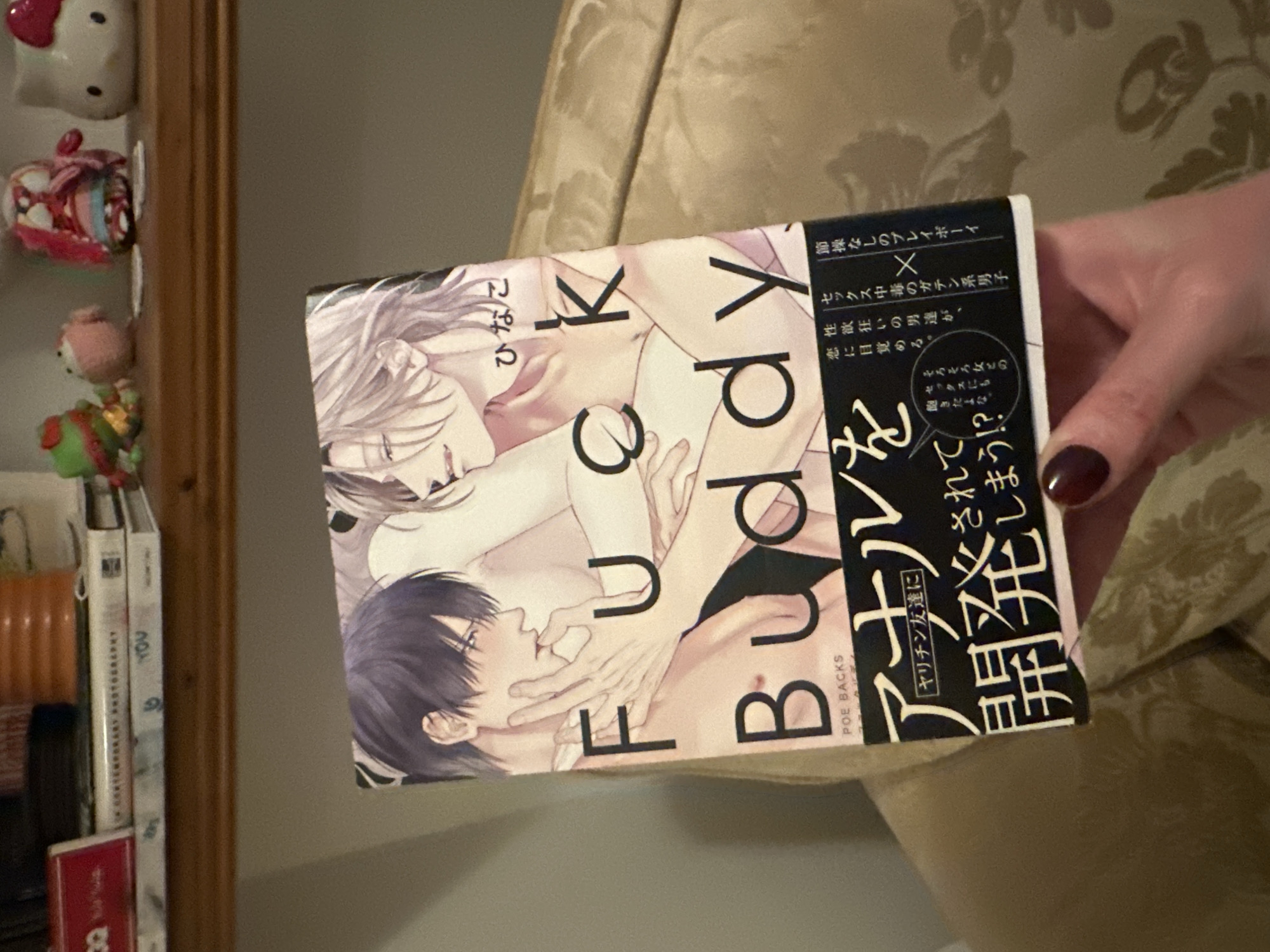 A hand holds the cover of an illustrated book titled "Fuck Buddy" against a textured background. The cover features two male figures in an intimate embrace. The book's spine displays Japanese characters. To the left, a shelf with decorative items and books is partially visible, including a Hello Kitty figurine and books titled "Contemporary Photography" and "YOU."