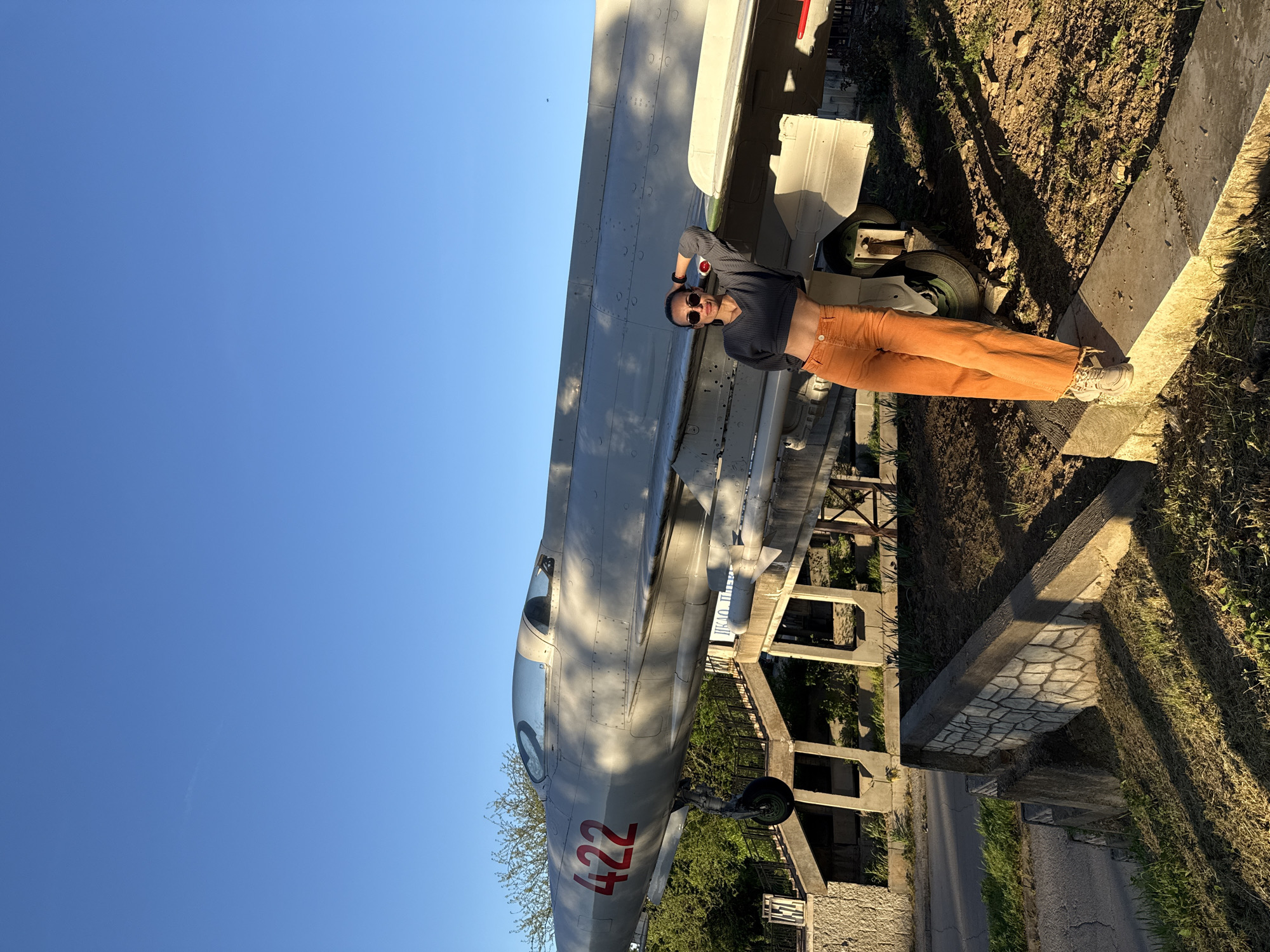 The image displays a person posing beside a vintage military jet under a clear, late afternoon sky in what appears to be an outdoor museum or monument in Pleven, Bulgaria.
The main subject is a person, likely a woman, wearing sunglasses, a dark grey long-sleeved crop top, and bright orange high-waisted pants with white sneakers. Her midriff is exposed, and she has a watch on her left wrist. She stands to the right of the airplane, looking directly at the viewer with one hand behind her head, striking a relaxed pose.
The dominant object is a light grey or silver vintage military aircraft, likely a MiG fighter jet, positioned horizontally. The prominent number "422" is stenciled in red on its fuselage near the cockpit. A missile or external fuel tank is visible beneath its wing, and its landing gear is extended.
The setting is an open-air environment with a clear, bright blue sky. The warm, low-angle sunlight of the golden hour casts long, distinct shadows across the ground and structures. The terrain consists of dry, earthy patches mixed with grass. To the right of the person, there's a concrete ledge or wall, and beyond it, an asphalt road section. In the background, concrete structures, possibly remnants or parts of a building or monument, are visible, partially obscured by green foliage. The overall scene suggests a historical display.
The only clearly legible text is the red number "422" on the aircraft. Some distant, blurred Cyrillic text is visible on a sign or building, but is unreadable.