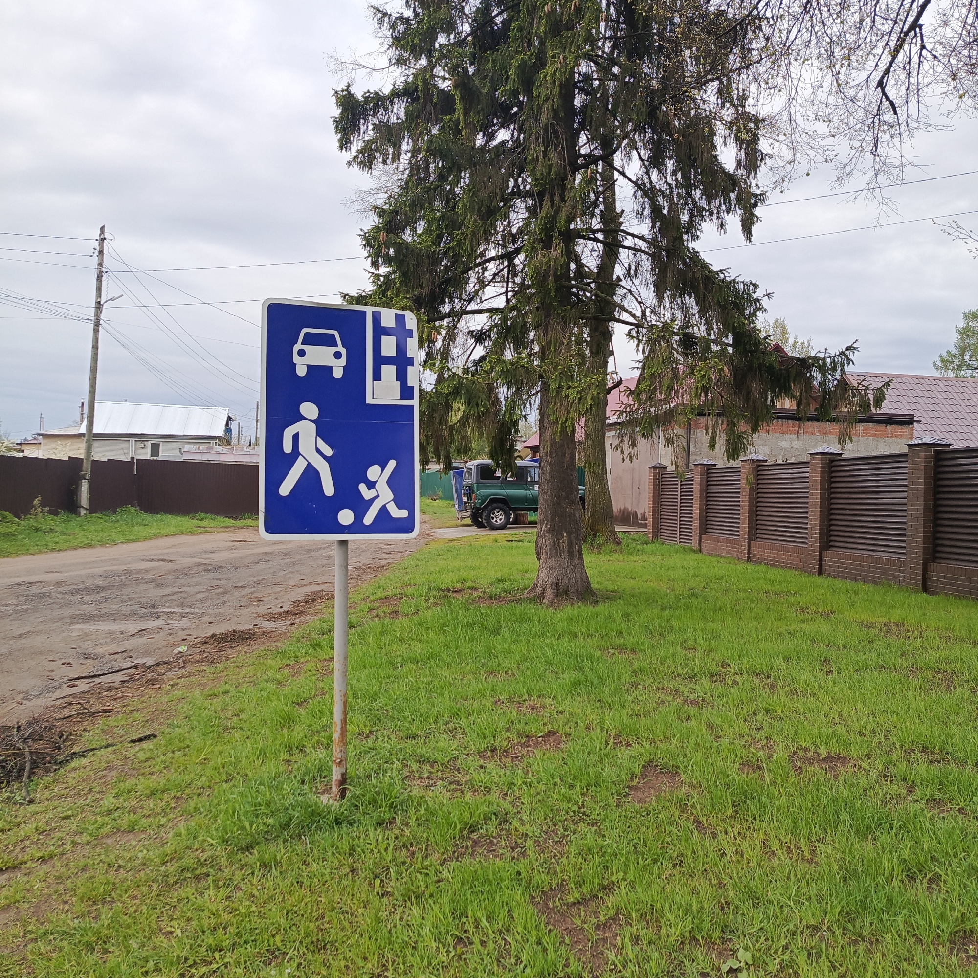 The image shows a traffic sign indicating a shared road for vehicles, pedestrians, and children at play. The sign is positioned on a metal pole on a grassy area adjacent to a cracked asphalt road. Behind the sign, there is a large evergreen tree and a section of a brown fence with brick columns. In the background, a green utility vehicle is parked near a house with a red roof, and a utility pole is visible on the left. The photo was taken in the city of Chaikovsky, Russia.