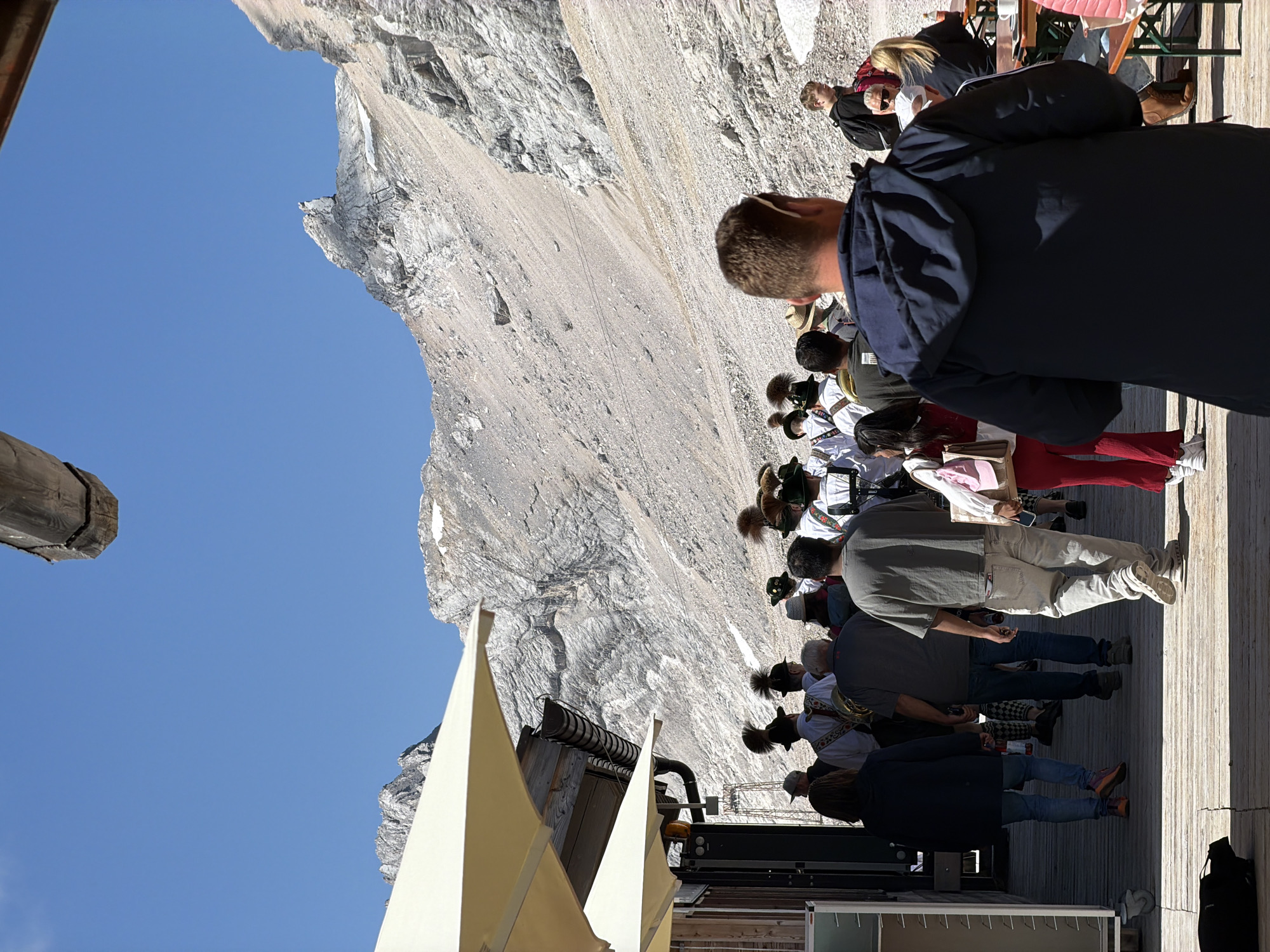The image captures a sunny day at a mountain resort, likely the Alpenrosensteig in Leutasch, Austria. In the foreground, a group of people are gathered on a wooden deck. To the left, a woman in a dark blue jacket and jeans walks away from the camera, accompanied by a person in a grey t-shirt and jeans. Several individuals in traditional Austrian attire, including feathered hats and white shirts with embroidered vests, are visible further back, some holding musical instruments. A large, rugged mountain face dominates the background under a clear blue sky. Outdoor umbrellas provide shade on the deck. The scene appears to be a tourist area with a strong cultural element, possibly a performance or procession.
