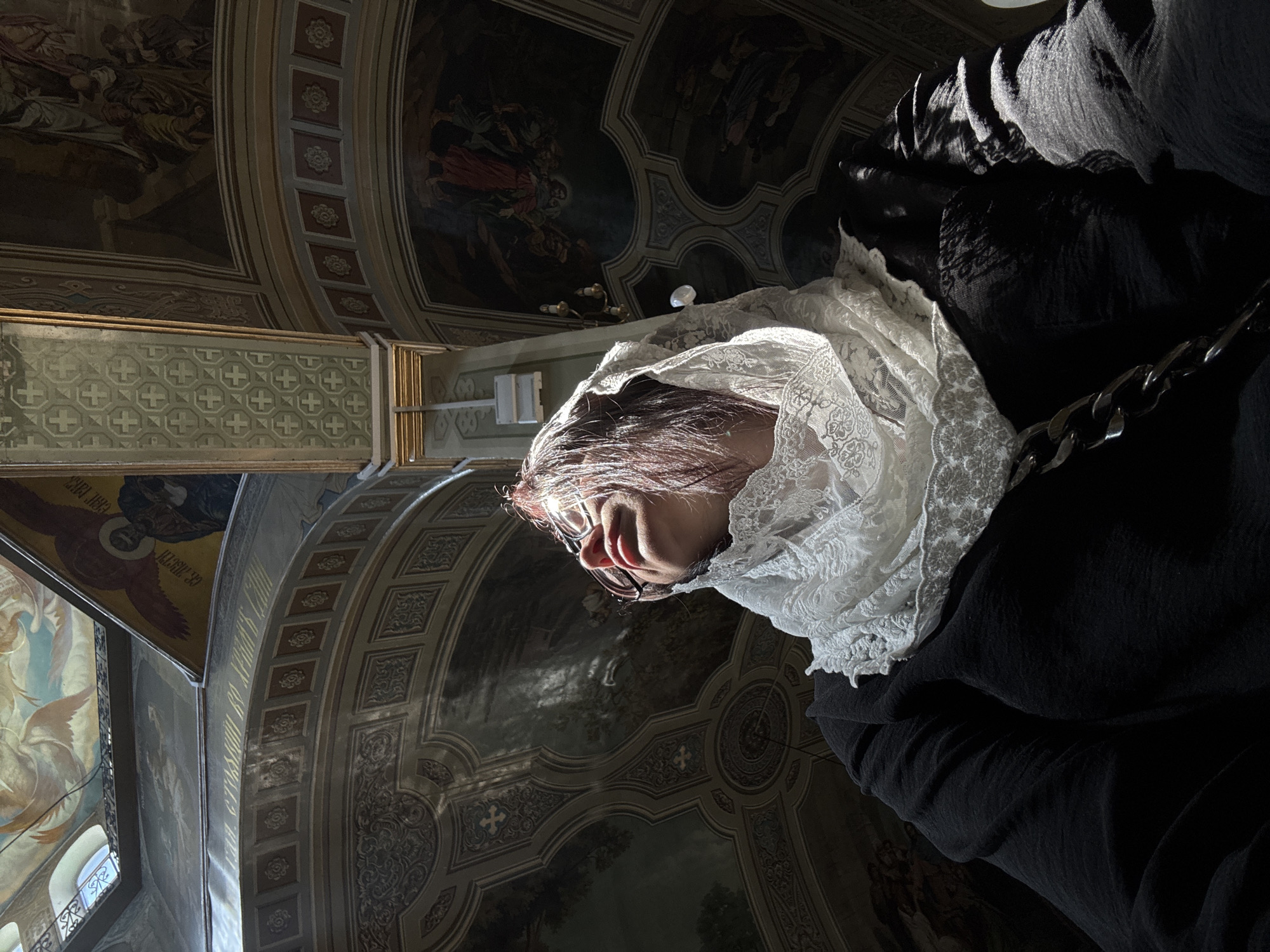 An indoor, low-angle shot captures a person wearing a white lace headscarf and glasses, looking upwards. The person is positioned in the foreground, with their face partially illuminated by sunlight. The background reveals the ornate ceiling of a church, adorned with frescos and intricate architectural details. The overall lighting is dramatic, with strong shadows and highlights, suggesting the presence of natural light filtering in from a window or opening. The environment appears to be a place of worship or historical significance, given the detailed religious artwork on the ceiling.