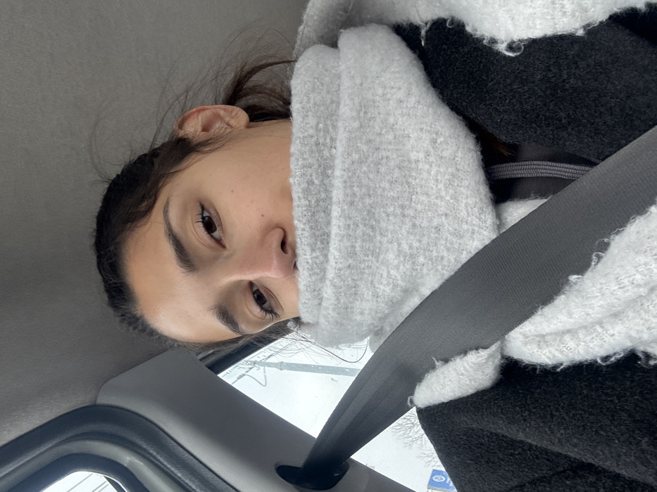 This image depicts a female subject, visible from the upper chest to the top of her head, seated inside a vehicle in Cibukovac, Serbia. Her dark hair is casually styled, and her light brown eyes are directed towards the camera. A prominent, light grey, chunky knit scarf covers the lower half of her face, from just below the nose, extending around her neck. She wears a dark-colored, likely black, coat, and a grey drawstring from an underlying garment is visible between the scarf and the coat. A black seatbelt is engaged, crossing her left shoulder and torso diagonally. The vehicle's interior is characterized by a grey headliner and a portion of a grey side pillar. Through the vehicle's window on the left, a blurred outdoor environment is visible, suggesting an overcast day with indistinct shapes of bare tree branches and a distant, unreadable blue and white sign.