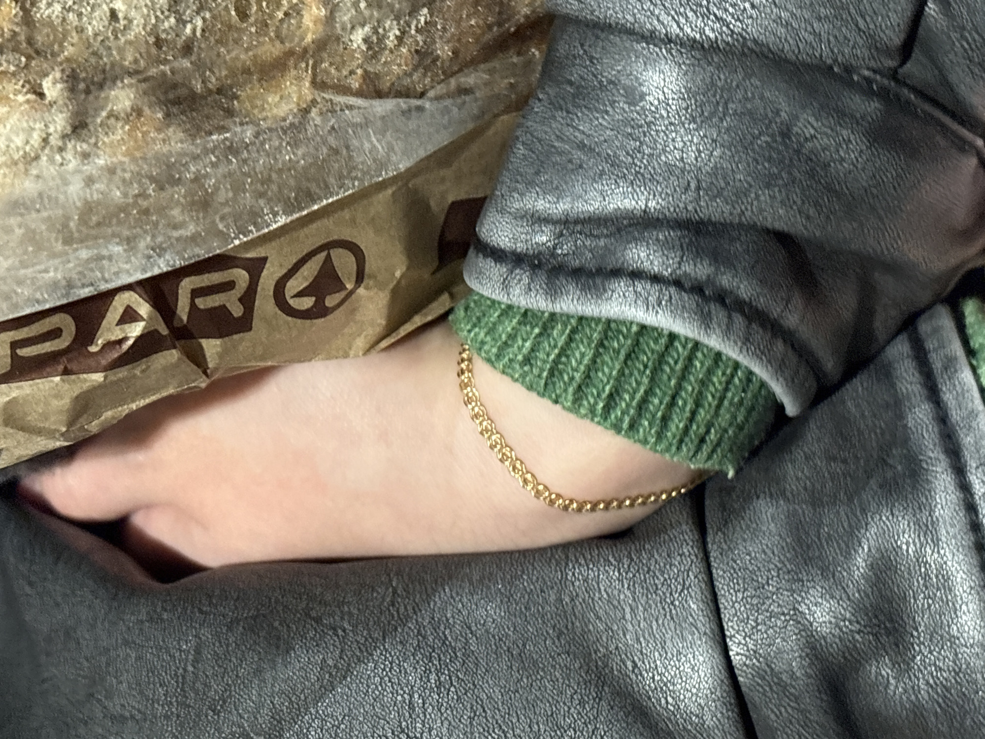 The image presents a close-up view of a person's arm resting against a grey leather jacket and a brown Spar grocery bag. The arm is adorned with a delicate gold chain bracelet. The green ribbed cuff of a sweater peeks out from beneath the jacket sleeve. Part of a baguette is visible above the Spar bag. The image provides no direct information about the location, but the presence of a Spar grocery bag indicates a location where the chain operates. As the prompt asks for the location in Kazan, Russia, it can be concluded that the scene takes place there.