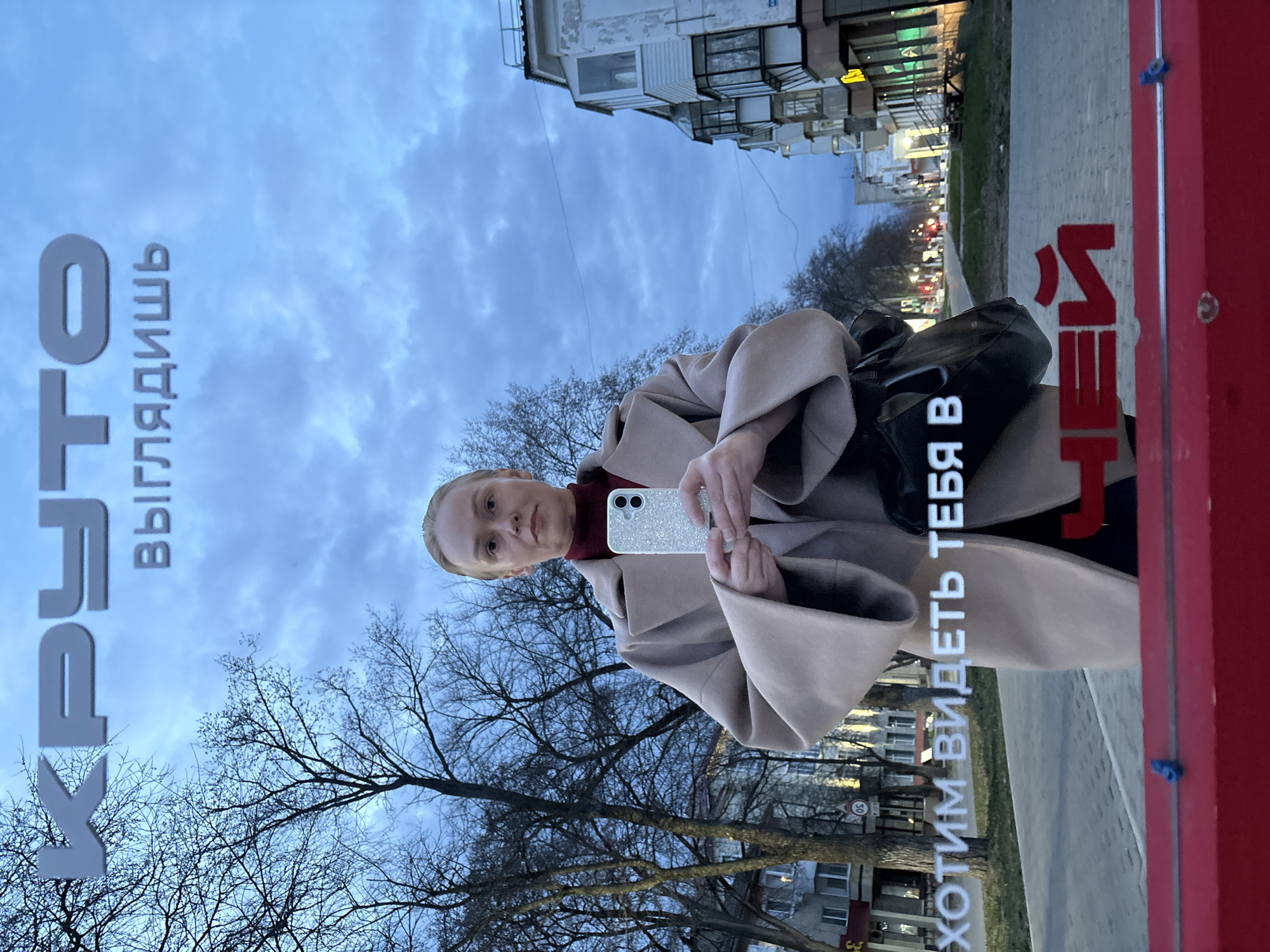 The image captures a reflection of a woman taking a selfie, likely off a large glass or mirrored surface, such as a shop window or a public information screen. The woman, with light hair pulled back, is wearing a red turtleneck or collar under a light beige or tan coat. She holds a silver or sparkly smartphone with a multi-lens camera system, pointing it towards herself. A black shoulder bag is visible on her left side.
The setting reflected behind her is an urban street in Chaikovsky, Russia, characterized by bare-branched trees, multi-story residential or commercial buildings, and a paved sidewalk. The sky is overcast with soft, diffused clouds, suggesting either late afternoon or early evening, a time further indicated by visible streetlights and traffic lights illuminated in the distance.
Visible in the reflection are parts of a red structure on the right, possibly an advertisement stand or bus stop. Russian text, mirrored due to the reflection, is prominently displayed vertically on both sides of the woman. On the left, the text reads "КРАТО ВЫГЛЯДИШЬ" (Krato Vyglyadish), which is very likely a misspelling or stylized rendition of "КРУТО ВЫГЛЯДИШЬ" (Kru-to Vyglyadish), meaning "You look cool/great." On the right, the text reads "ХОТИМ ВИДЕТЬ ТЕБЯ В НЕЙ" (Khotim videt' tebya v ney), translating to "We want to see you in her/it," suggesting an advertisement for clothing or style. The woman's expression is neutral as she looks directly at her phone's camera.