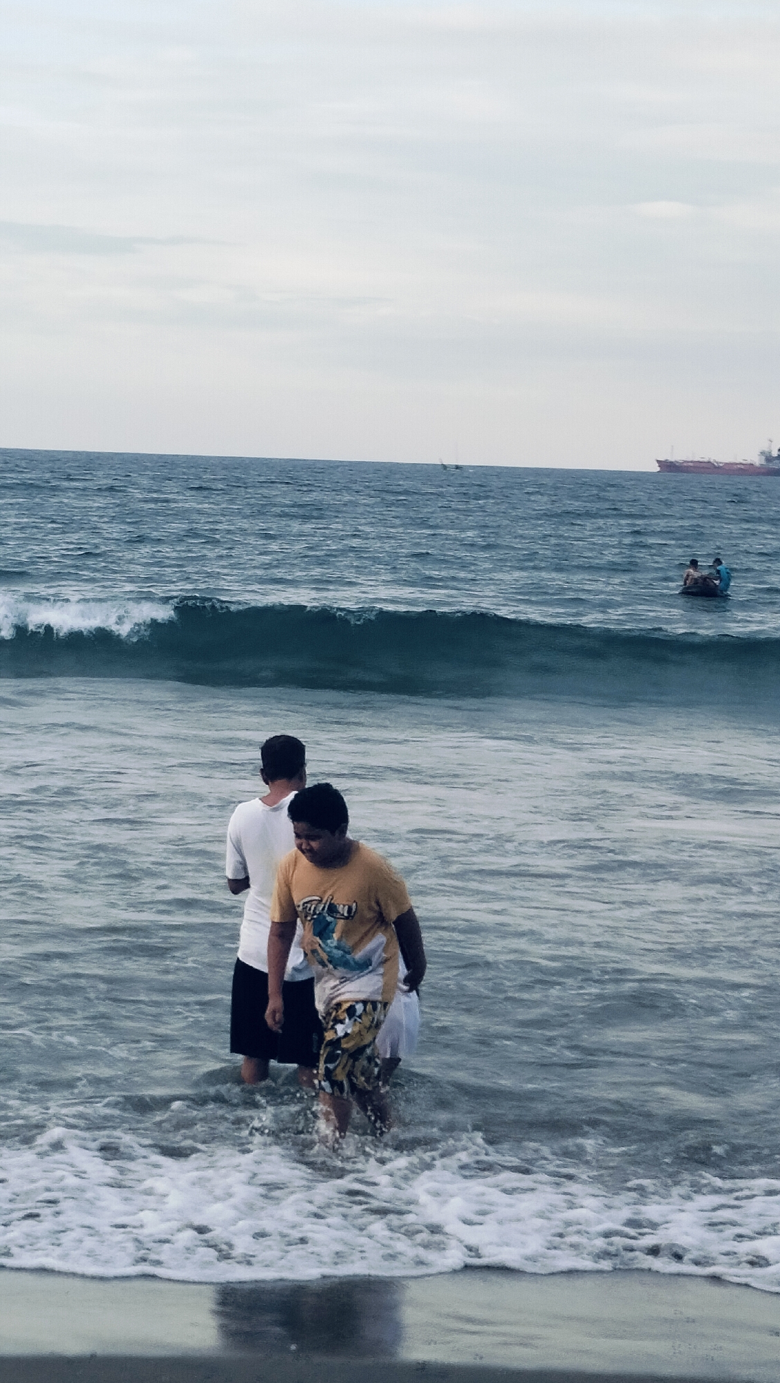 This image depicts a coastal scene in Lhokseumawe, Indonesia, on an overcast day. The foreground shows two individuals partially submerged in shallow ocean water. The person on the right, closer to the camera, is a male wearing a yellow t-shirt with a graphic design and patterned shorts, walking towards the viewer with water reaching his mid-thigh. Behind and to the left of him, another male is visible from the back, wearing a white t-shirt and dark shorts, facing the sea with water around his waist. White foam from breaking waves is visible around their feet.
In the midground, a prominent wave is breaking, stretching across the frame. Further out in the water, two more individuals are observed floating on what appears to be a dark, low-profile craft or inflatable object. One wears a blue top, and the other a lighter-colored top.
The background features the expansive ocean under a light gray, cloudy sky. On the far left horizon, a thin, vertical structure resembling a mast is visible, suggesting a distant small vessel. On the far right horizon, a large red cargo ship or tanker is clearly discernible. The overall impression is one of people engaging with the ocean under subdued lighting conditions.