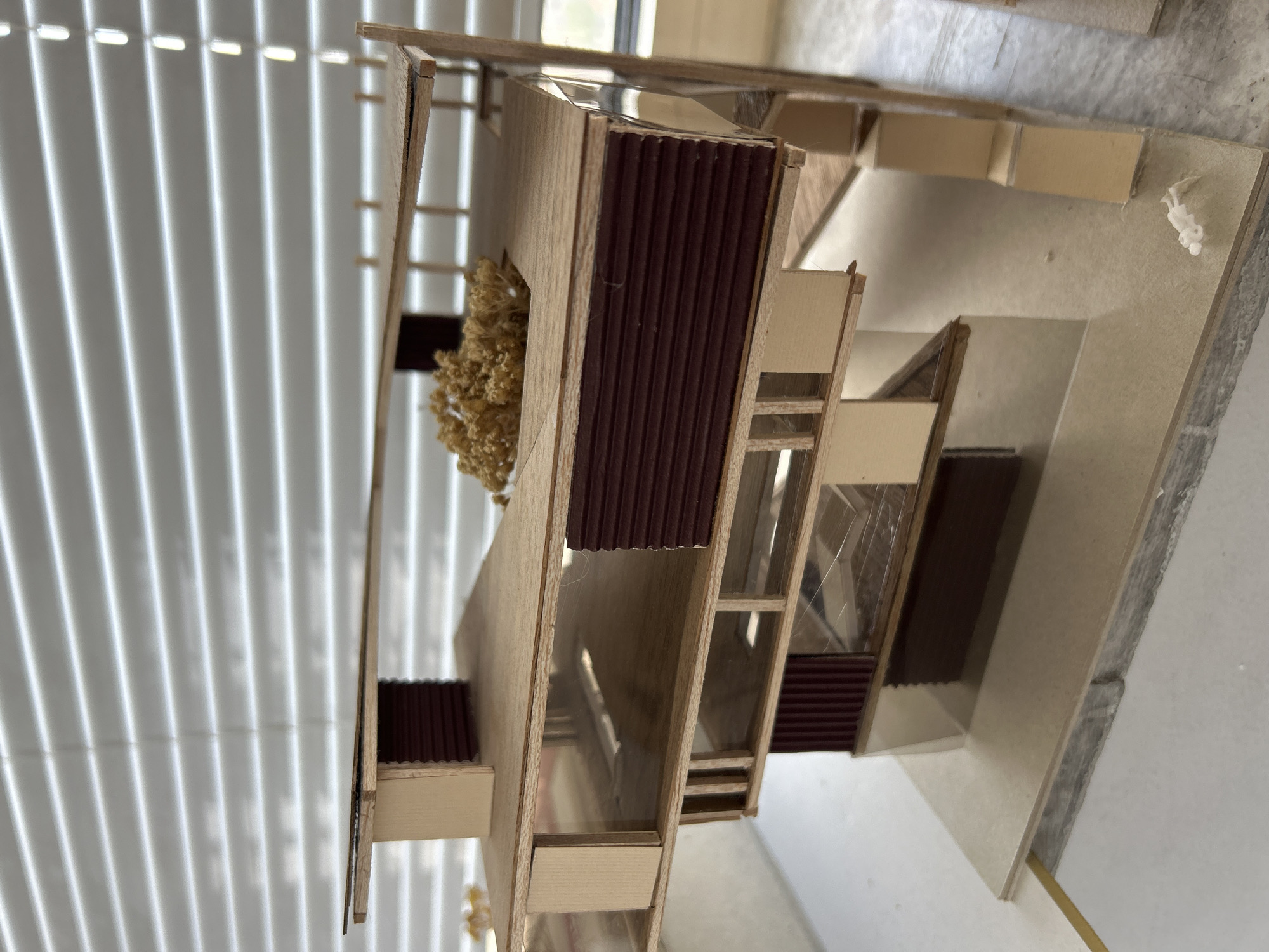 The image shows a scale model of a building with a modern design, likely an architectural model. The model is constructed from light-colored wood and features elements like vertically ribbed brown panels, simulated windows with clear material, and a small artificial tree. The base of the model appears to be made of a similar material to the building's construction, with light beige/grayish color. The backdrop includes white blinds, suggesting an indoor environment. The location is not possible to determine using the image.