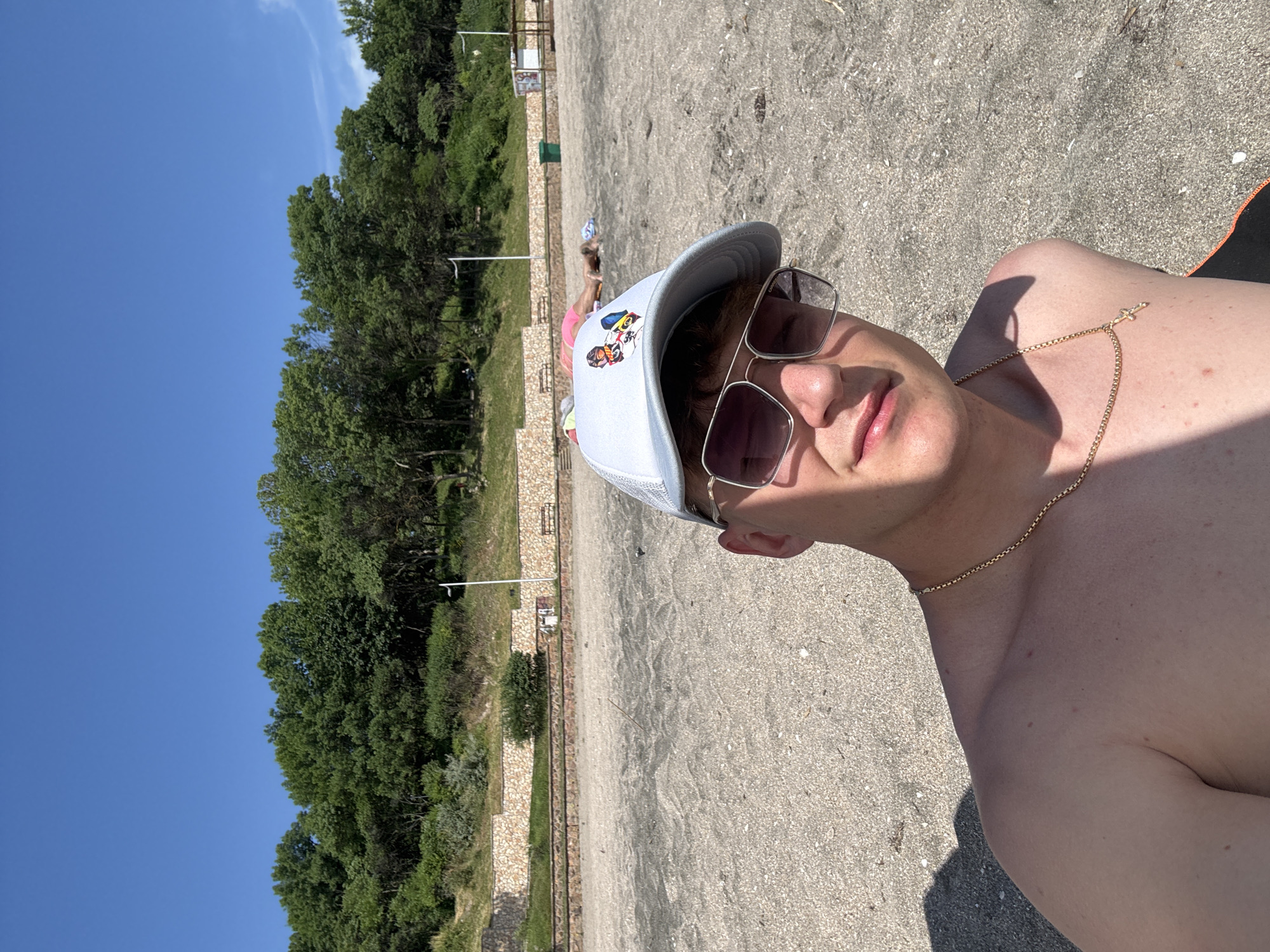 A young Caucasian male is taking a selfie on a sandy beach. He is wearing a light-colored baseball cap and aviator sunglasses, and a gold chain with a cross pendant. His bare shoulders and upper chest are visible. The background shows a sandy beach with some scattered debris, followed by a stone wall and a line of trees. Several other beachgoers are visible in the distance, lying on towels. The sky is clear and blue. The location is the sandy beach area of Burgas, Bulgaria.