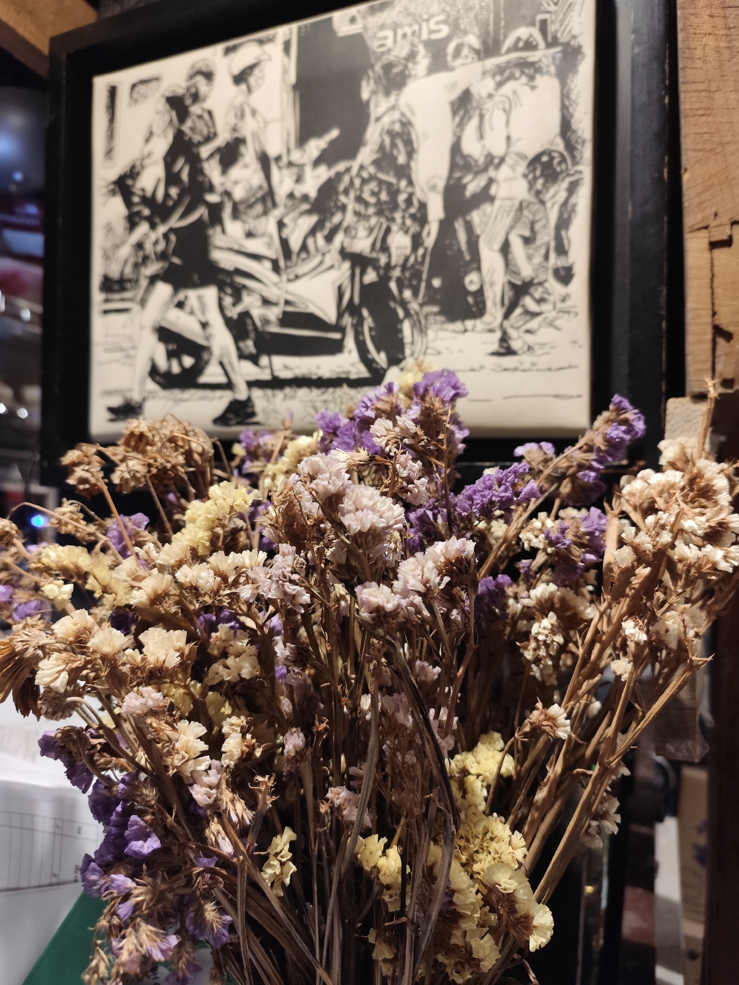 A close-up shot captures a bouquet of dried flowers in the foreground, featuring muted shades of purple, cream, and pale yellow, along with their brown, brittle stems. The flowers are in sharp focus, creating a delicate texture that dominates the lower and central part of the frame.
In the blurred background, a framed, black-and-white illustration or artwork is visible. This artwork depicts several stylized figures, appearing to be people, some in dynamic poses near or interacting with motorcycles or scooters. On the left side of the artwork, a figure in a dark top and skirt stands beside a scooter. Towards the right, a group of figures includes one wearing a helmet, another bending, and a small child-like figure kneeling. The style suggests a comic book or graphic novel aesthetic, portraying an outdoor, possibly urban, scene. The word "amis" is discernible in the upper right section of the artwork.
The setting appears to be indoors, possibly a cafe or a cozy establishment, indicated by the artwork and rustic wooden elements visible on the right side of the frame. The lighting is ambient, without clear indications of the time of day. No direct activity is occurring in the captured scene, only the static arrangement of flowers and the background illustration. While the location context of Da Lat, Vietnam is provided, no specific visual cues within the image confirm this.