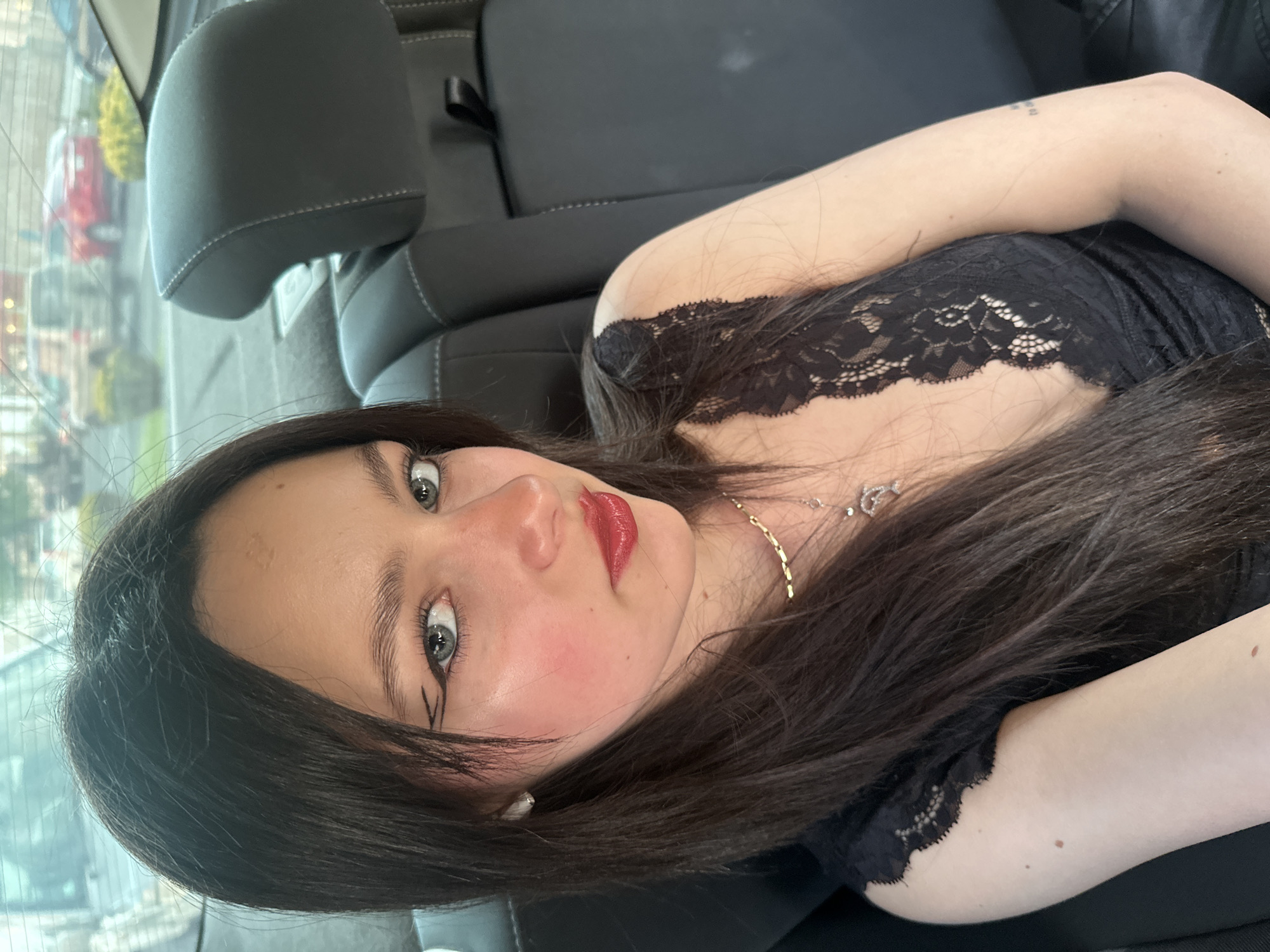The image shows a woman with long dark hair, red lipstick, and pale skin, wearing a black lace top. She is lying in the back seat of a car. The headrest of the car seat is visible behind her head. Through the car window, there is a blurred cityscape with cars and buildings, suggesting the image was taken in an urban environment. It's not possible to determine the specific location in Yerevan, Armenia.