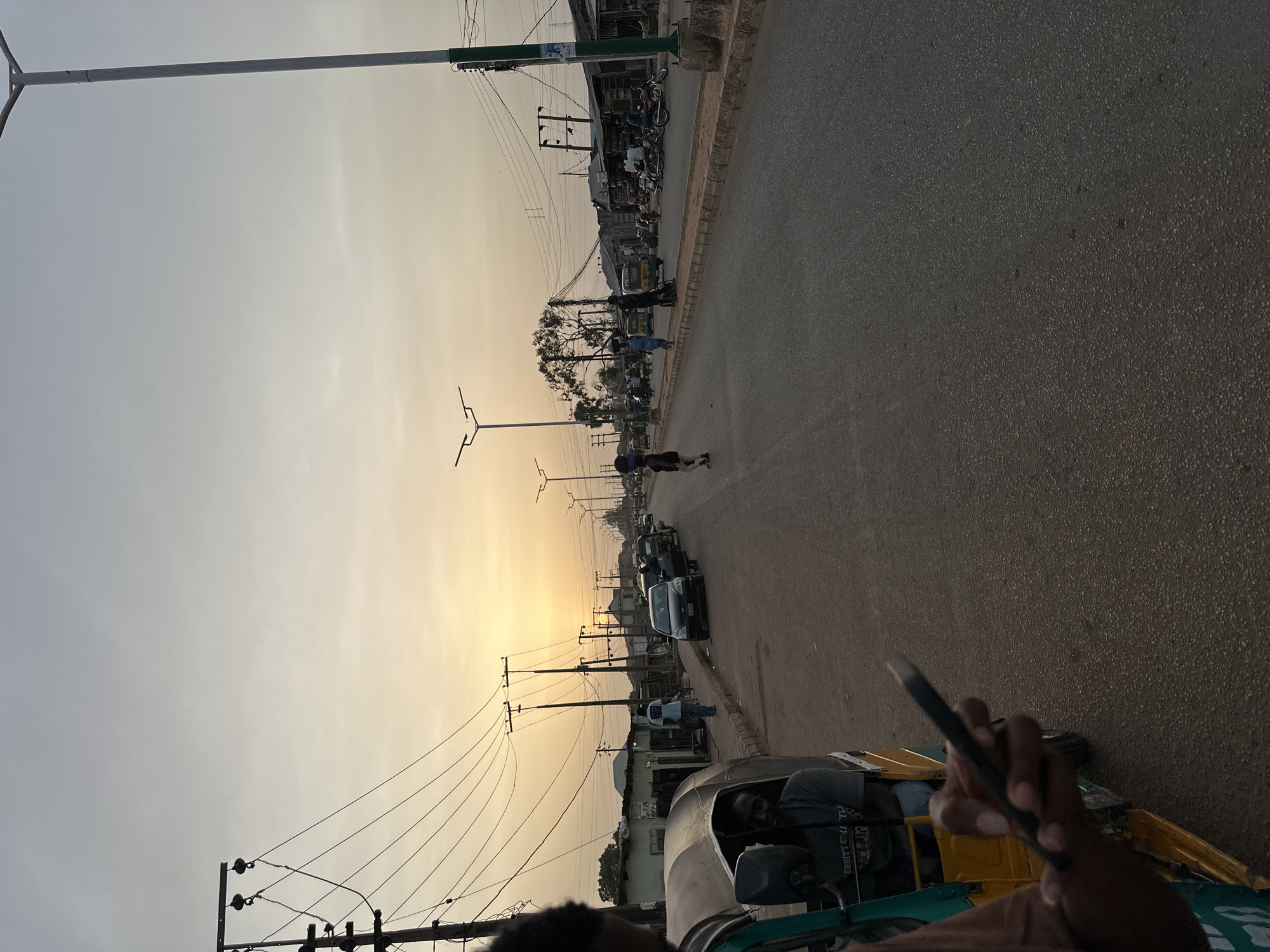 This media file captures a street scene in Bukuru, Nigeria, during what appears to be the late afternoon or early evening, evidenced by the warm, golden light on the horizon. The image is taken from a low perspective, likely from inside a moving vehicle, with a hand holding a smartphone visible in the foreground (bottom right when correctly oriented). The vehicle seems to be a motorized tricycle, commonly known as a "keke napep," with its driver partially visible.
The scene depicts a bustling urban environment. An asphalt road stretches into the distance, flanked by a sidewalk and various low-rise buildings, likely shops or residences, on the right. Numerous utility poles with a dense network of overhead wires line the street, a common sight in the region. Several streetlights are also visible along the road.
People are seen walking on the sidewalks and crossing the street. Various vehicles, including motorcycles, cars, and other tricycles, are parked or moving along the road. One person is distinctively walking in the middle of the road in the mid-ground. The sky above transitions from a light, hazy grey-blue overhead to the warm, yellowish glow near the horizon, suggesting the approach of sunset or sunrise, though the activity level points more towards evening.
Notable details include the specific type of informal public transport (keke napep) and the visible infrastructure. On the dark shirt of the person inside the foreground tricycle, partial text can be seen, reading "UNIVERSITY GRA...".