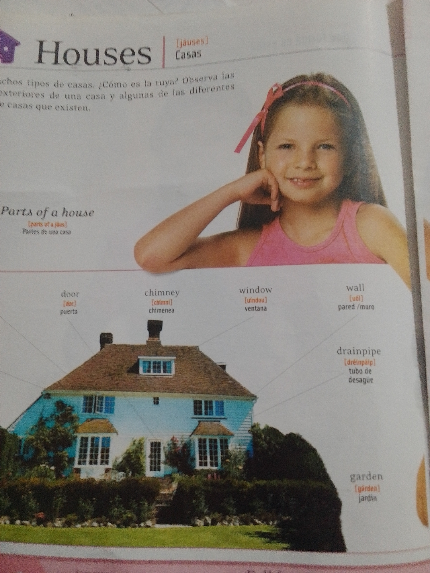 This is a book page with an illustration of a light blue house and its parts. The title "Houses" is prominently displayed at the top of the page, with "Casas" in parentheses. Below the title, there is a paragraph in Spanish discussing different types of houses. A young girl with a pink headband and bow is pictured in the upper right corner, leaning her head on her hand and smiling. The page also details various parts of a house with their English and phonetic spellings, as well as their Spanish translations. These include "door" (puerta), "chimney" (chimenea), "window" (ventana), "wall" (pared/muro), "drainpipe" (tubo de desagüe), and "garden" (jardín). The house in the illustration has a brown tiled roof, several windows, and is surrounded by lush greenery, suggesting a pleasant, possibly daytime setting. The overall tone of the page is educational, aimed at teaching about houses and their components. The location context provided is La Tagua, Venezuela, though this information is not visually present in the image itself.