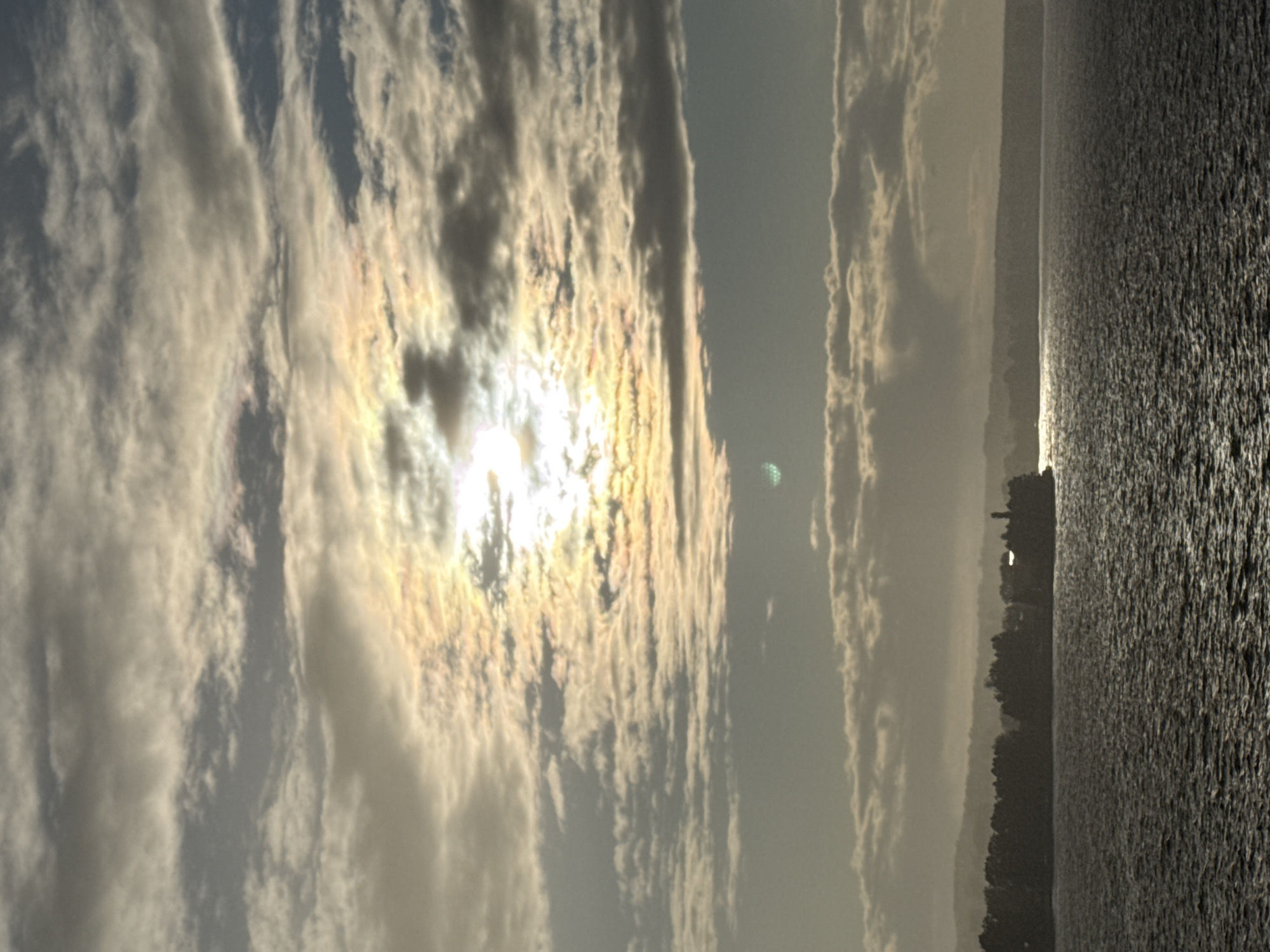 The image depicts a body of water, likely a river or lake, with a shoreline featuring a dense line of trees and some structures, including what appears to be a lighthouse or tower. The water's surface is textured with ripples, reflecting the subdued, hazy light of the sky. The sky itself is filled with clouds, some of which are tinged with the warm hues of sunlight breaking through. The overall atmosphere suggests a cool, possibly overcast day. This scene is situated in Votkinsk, Russia, a city known for its scenic waterfront.