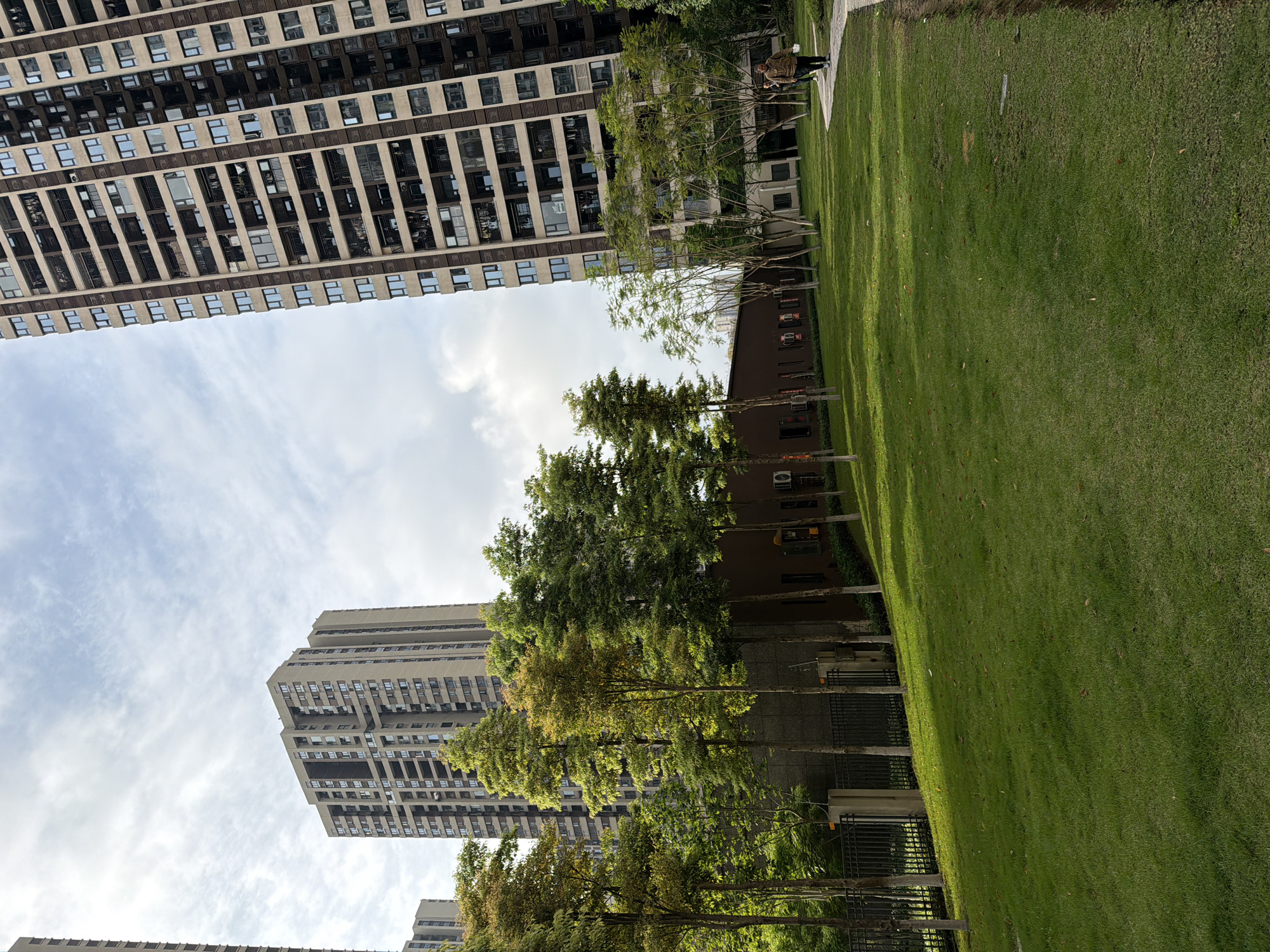 The image captures a vibrant urban residential scene in Chongqing High-tech Zone, China. A prominent high-rise apartment building, constructed with a light-colored facade and featuring numerous windows and balconies, dominates the left side of the frame, reaching towards a partly cloudy sky.
On the right, a vast, well-maintained green lawn stretches across the foreground and middle ground, bathed in sunlight that casts shadows from nearby trees. A paved pathway runs alongside the grassy area, where a single person is seen walking away from the viewer. The individual is dressed in a brown jacket and dark trousers, carrying a light-colored object.
A line of mature trees with lush green foliage, interspersed with some yellowing leaves, separates the lawn from a lower, dark-colored building in the mid-ground, which has visible air conditioning units on its exterior. Further in the background, another tall residential building is visible.
The time of day appears to be late morning or afternoon, given the clear sky and the angle of the sunlight creating distinct shadows. The overall setting suggests a modern and well-planned residential complex. No visible text is present in the image.