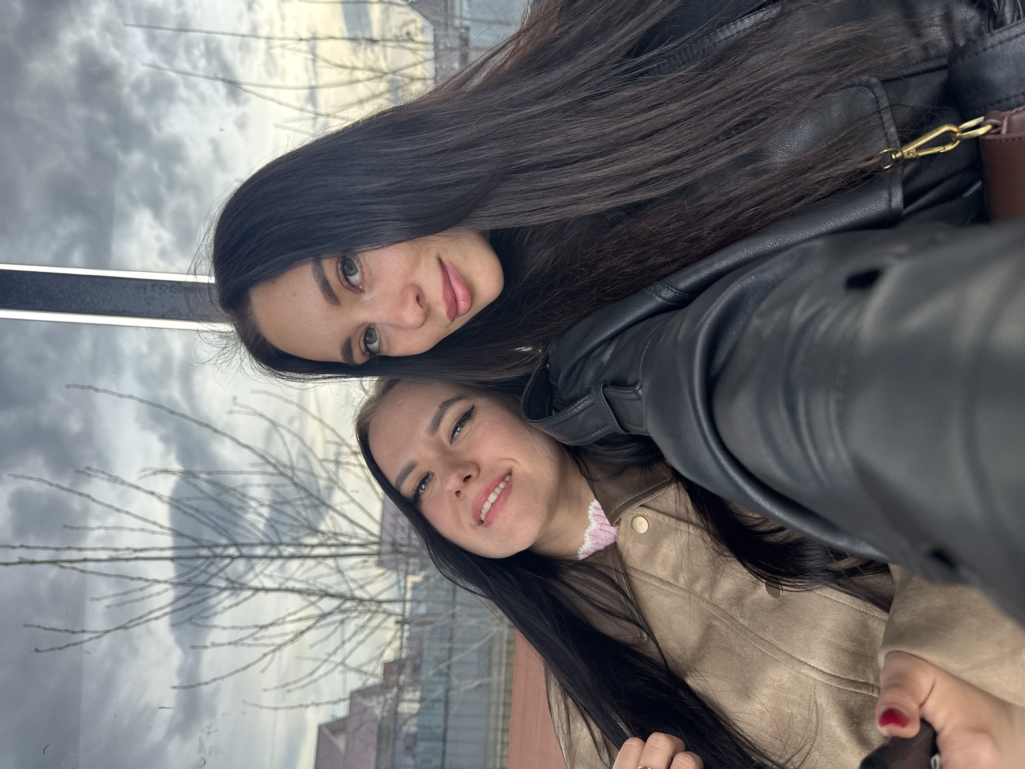 Two young women pose closely together for a selfie. The woman in the foreground wears a light brown jacket with gold buttons and a pink frilled collar peeking out from underneath. Her long, dark hair falls around her face and shoulders. She is smiling with slightly parted lips. The woman behind her wears a black leather jacket, and her long, dark hair cascades down. She also smiles at the camera. The background shows a cityscape reflected on a glass or window surface. Based on the architecture and details visible in the reflections, the location is likely Chaikovsky, Russia. The reflections include trees with bare branches and buildings with pink and gray tones, suggesting an urban environment.