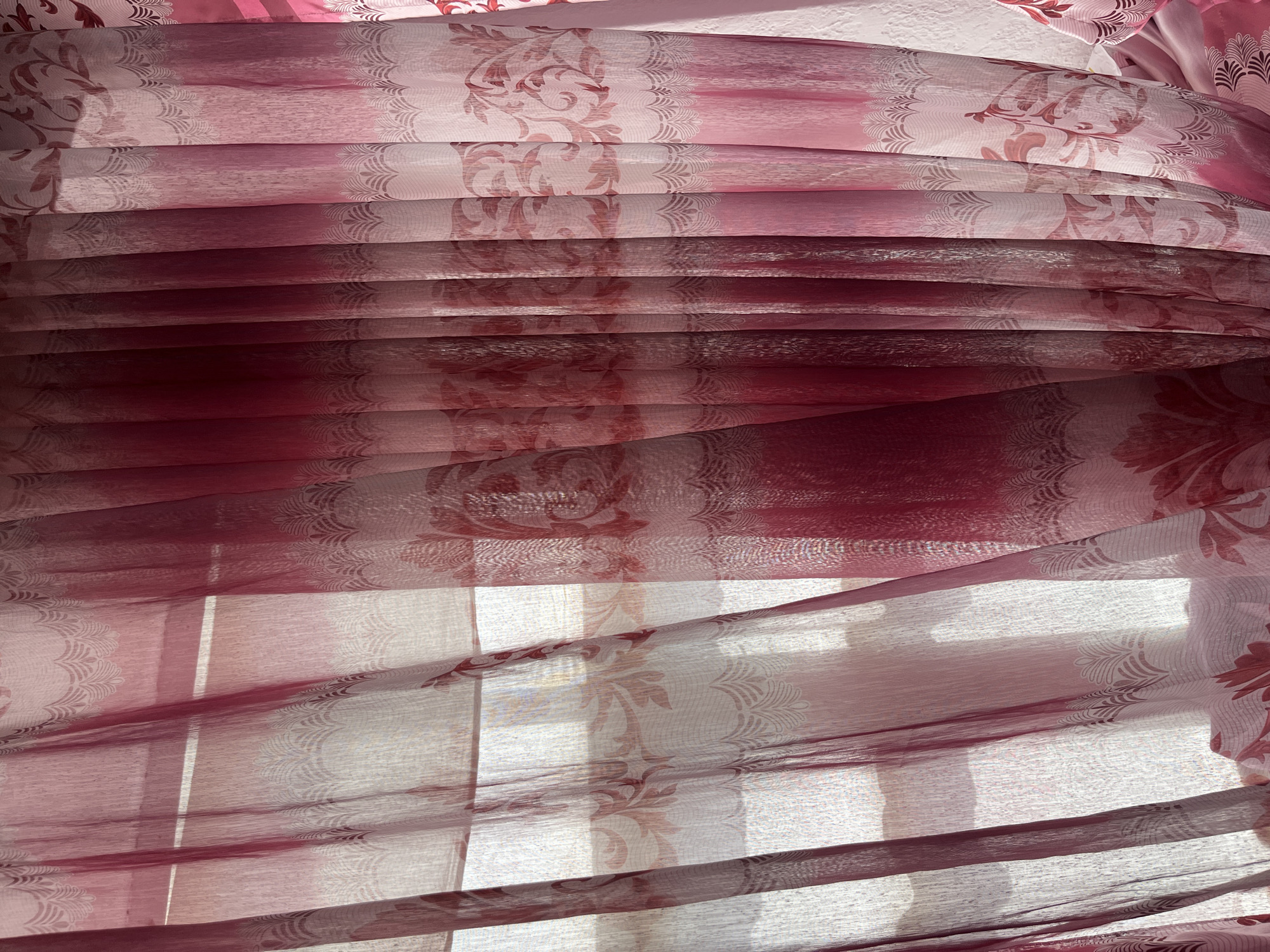 A close-up, eye-level shot captures a set of sheer, patterned curtains, predominantly in shades of pink and red, hanging in front of a window. The curtains are made of a translucent fabric, allowing diffused sunlight to filter through, illuminating the intricate floral and lace-like patterns that adorn them. The folds of the fabric create vertical lines and shadows, adding depth and texture to the image. The patterns include stylized floral motifs and delicate, fan-shaped designs along the bottom edges of some panels. The overall impression is one of soft light and decorative fabric. There are no people or discernible objects from the city of Batken City, Kyrgyzstan present in this image.