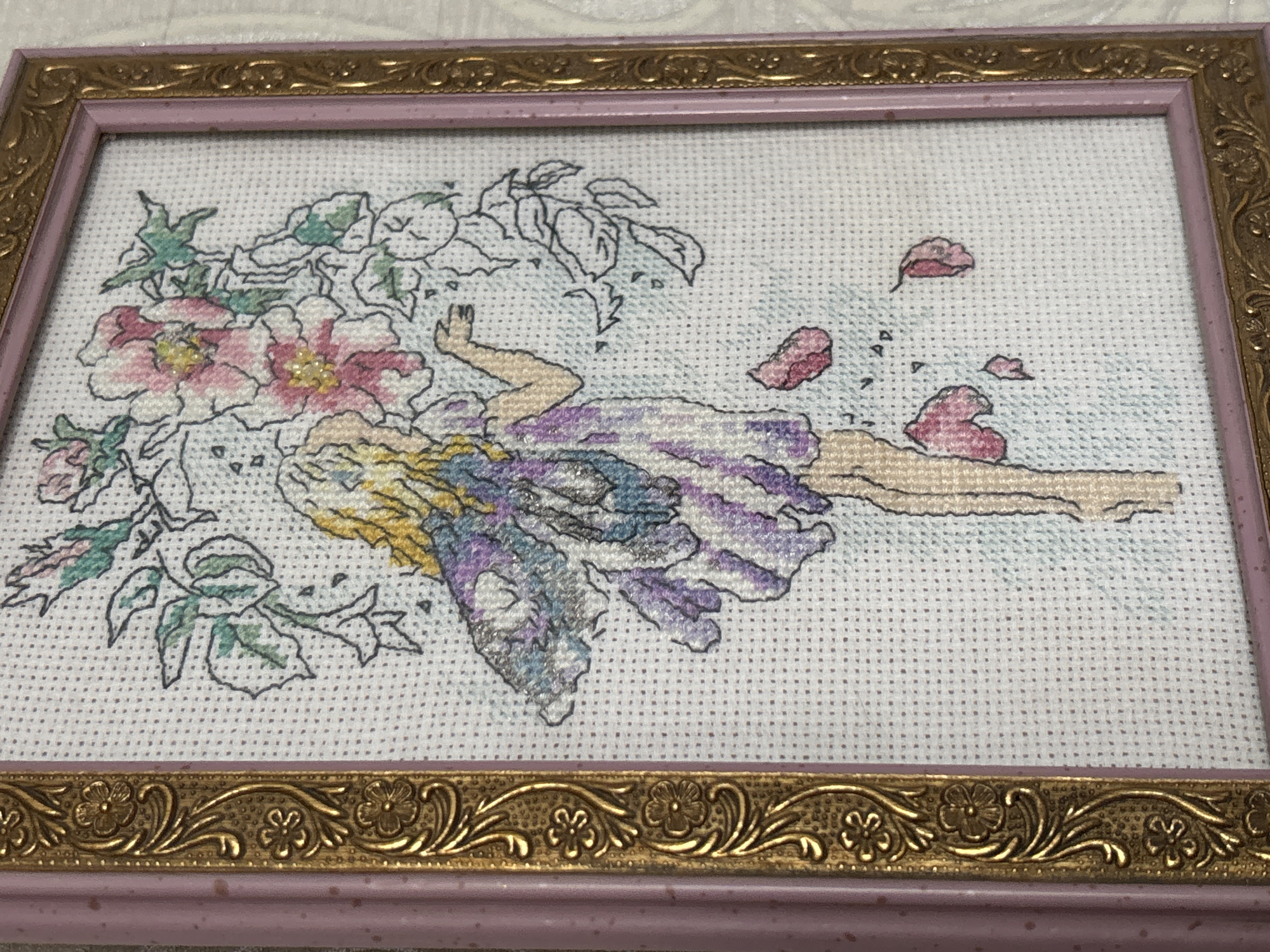 The image shows a framed cross-stitch artwork of a fairy standing among flowers. The fairy has blonde hair, purple and blue wings, and a striped purple dress. Pink and white flowers are depicted above and around the fairy, with some fallen petals scattered below. The cross-stitch is on white Aida cloth, and the frame is a light pink with ornate gold detailing on the inner border. The background behind the frame appears to be a textured, light-colored wall. The image is taken from a slightly elevated angle, looking down at the framed artwork. There is no visible information or context to suggest a location in Pavlodar, Kazakhstan.