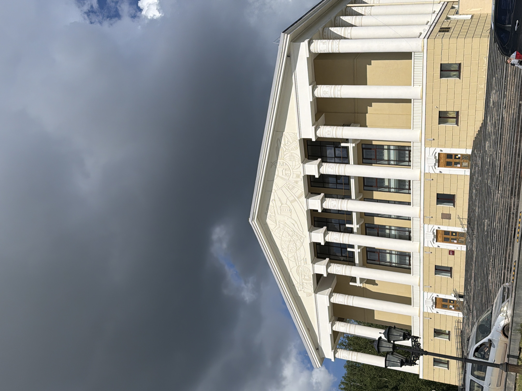 A low-angle, outdoor shot captures the front of a building with a Greek Revival architectural style. The building has white columns and a light yellow facade. Dark storm clouds fill a significant portion of the sky to the left of the building. Architectural details are visible on the building's pediment. Windows are regularly spaced on multiple floors. Stairs lead up to the building's entrance. Cars are parked near the building. A street lamp is near the base of the building.
Based on the architectural style, particularly the columns, and the distinctive mural-like design on the building's pediment, this is the Chaikovsky Music School in Votkinsk, Russia.