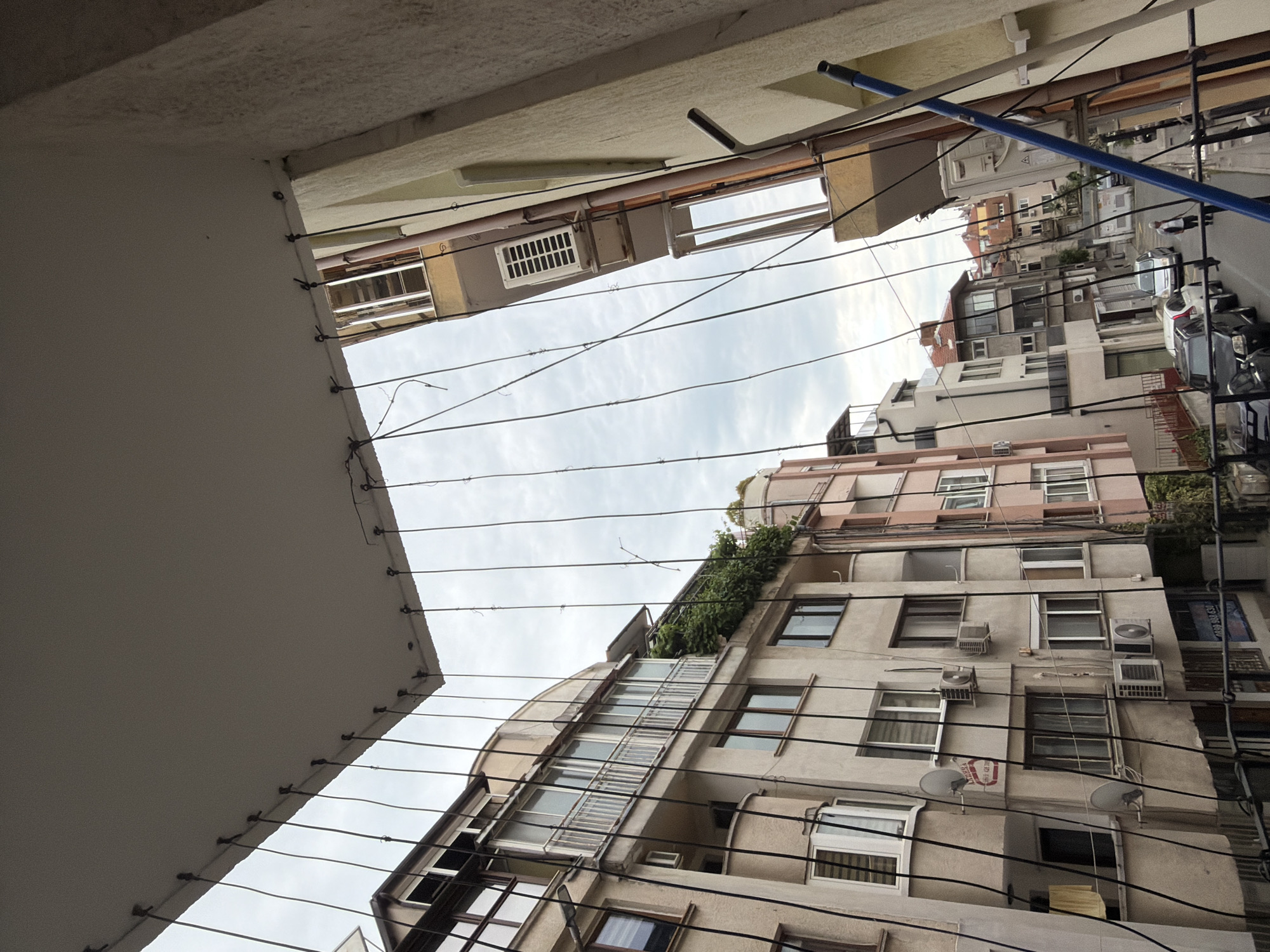 The image depicts an urban scene in Burgas, Bulgaria, shot from a low angle, looking up between buildings. Rows of clotheslines stretch across the frame, partially obscuring the view. The buildings are a mix of residential apartments, constructed with stone and beige concrete with white window frames and various types of air conditioning systems. There are buildings with large windows and balconies. Utility wires are strung across the sky. A blue laundry drying rack protrudes from a higher floor window. A street can be seen between the buildings, containing parked and moving vehicles along with pedestrians. The sky is overcast. The image captures the characteristic density and layered appearance of a central-European city.